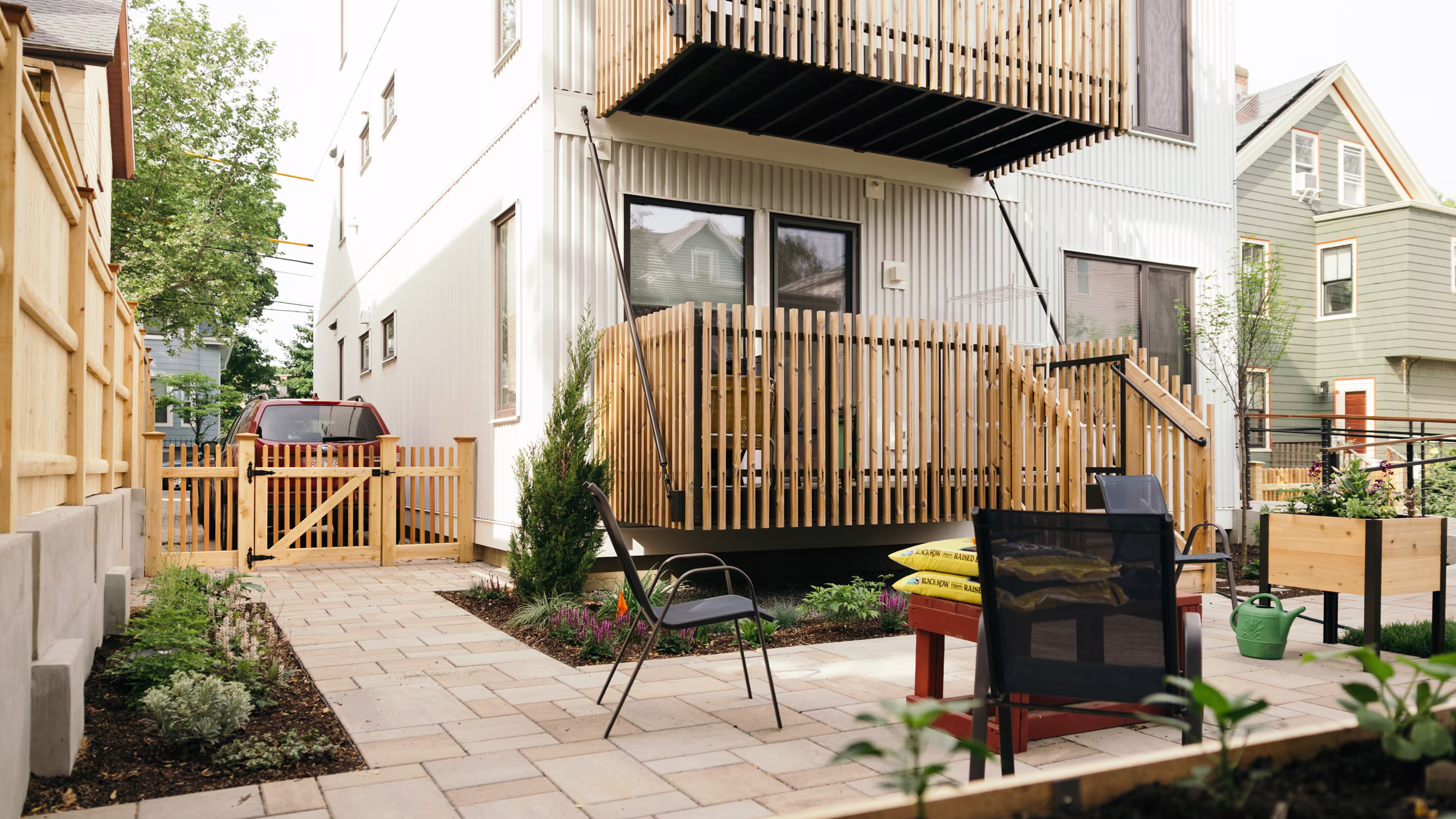 Backyard patio of a completed Reframe Systems multifamily building, with a wood slat deck, landscaped garden beds, paver walkway, and a wood gate opening to the street.