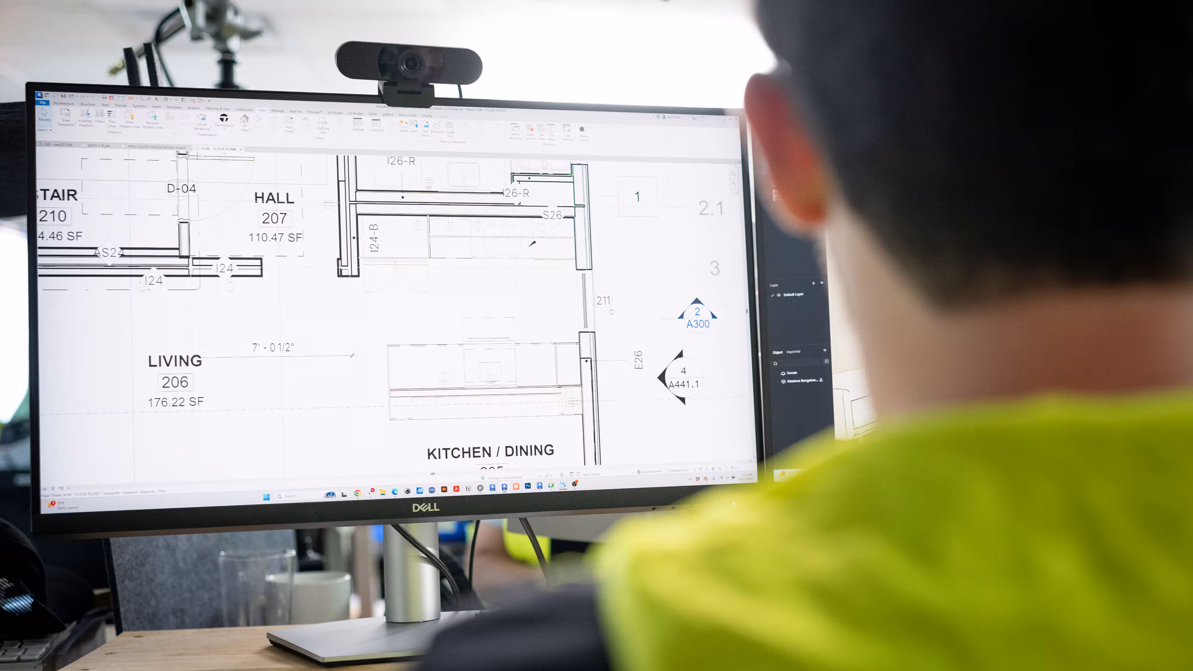 A Reframe Systems team member in a hi-vis vest reviews detailed modular home floor plans — including living, kitchen, dining, and hall spaces — in CAD software on a Dell monitor