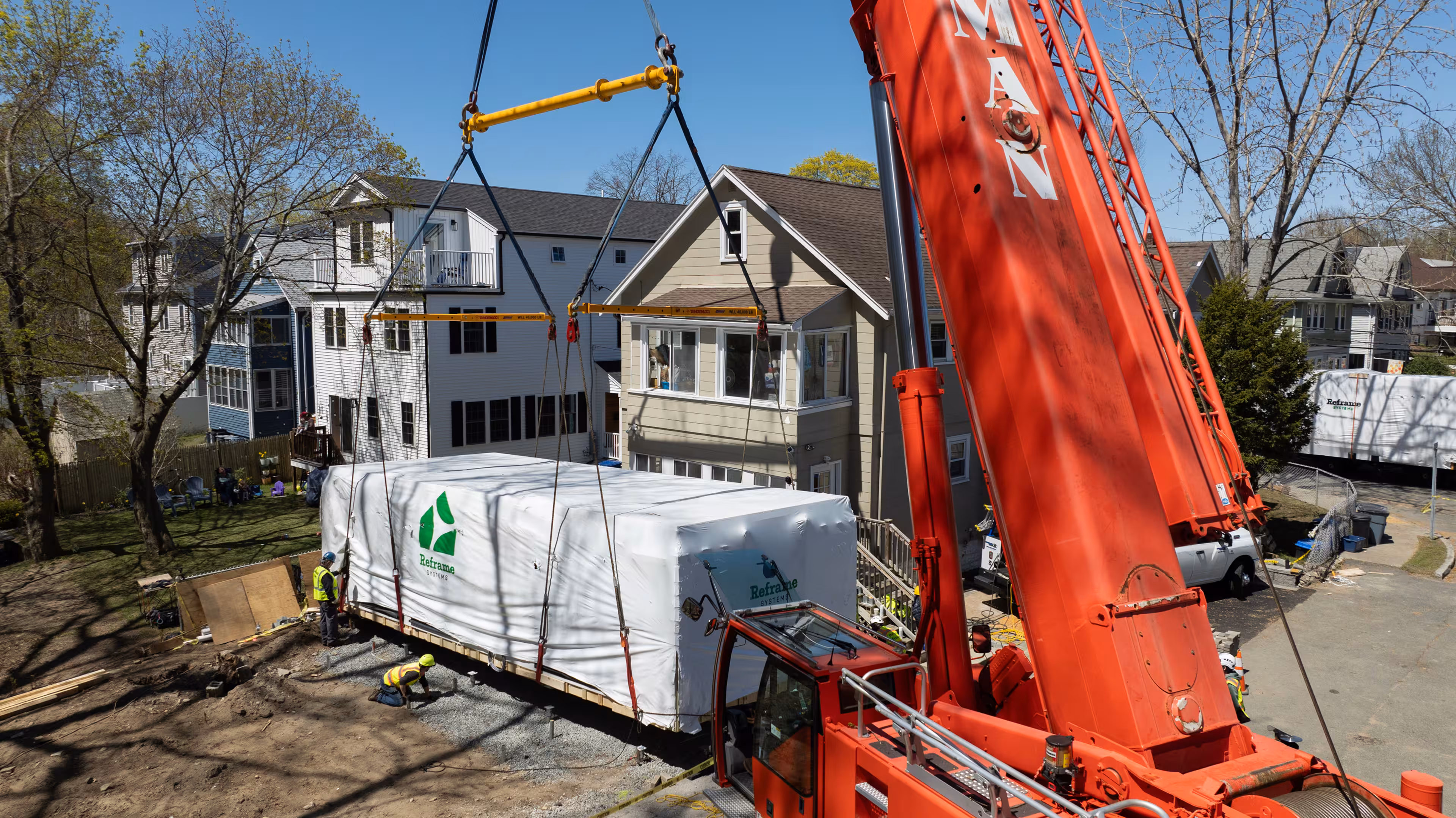 Aerial view of a crane lifting a wrapped Reframe Systems modular home module onto a residential job site in Devens, Massachusetts, with the installation crew on the ground below