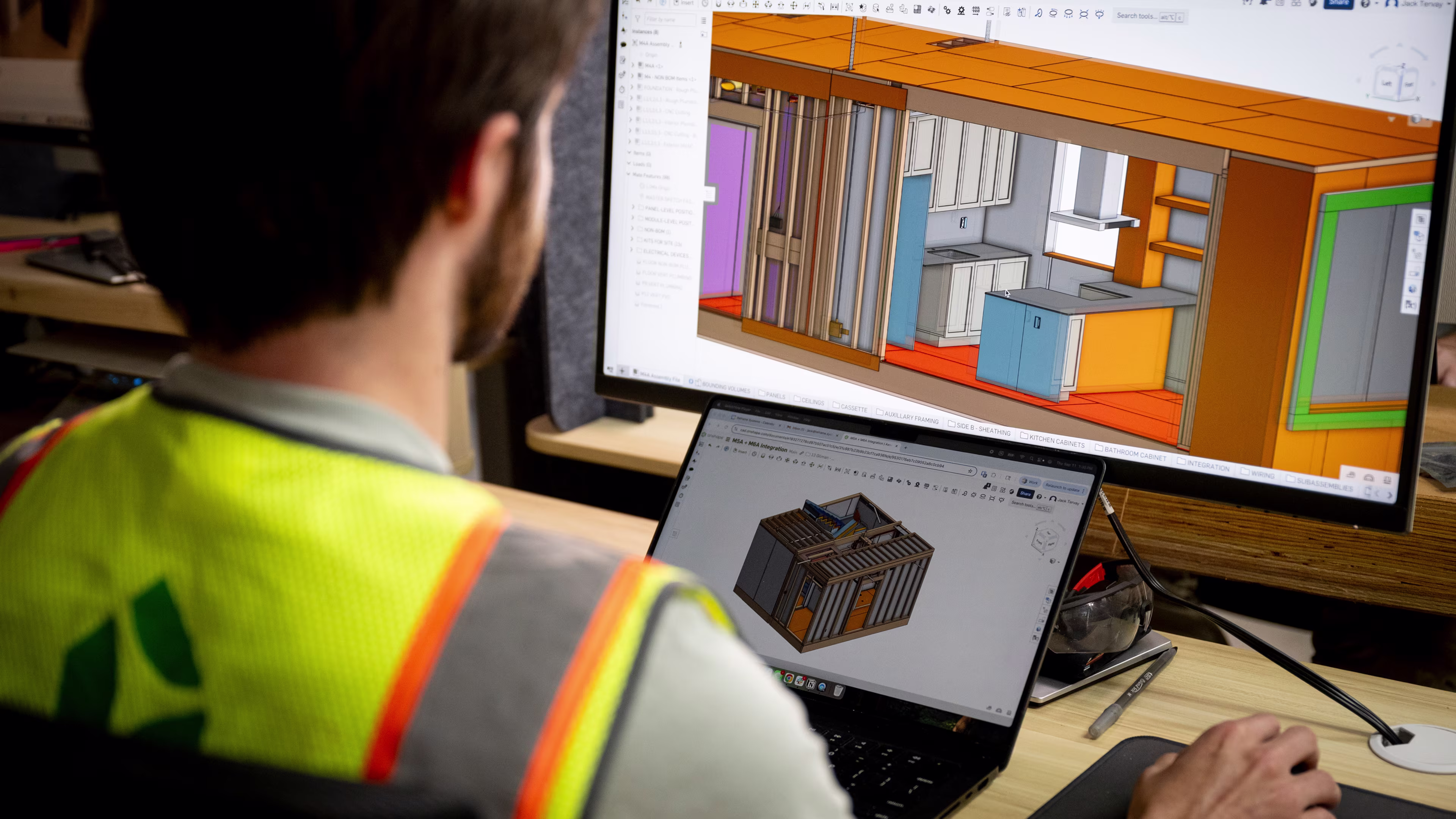 A Reframe Systems team member in a hi-vis vest reviews 3D CAD models of a modular home interior and structural module across a dual-screen workstation at the Andover production facility