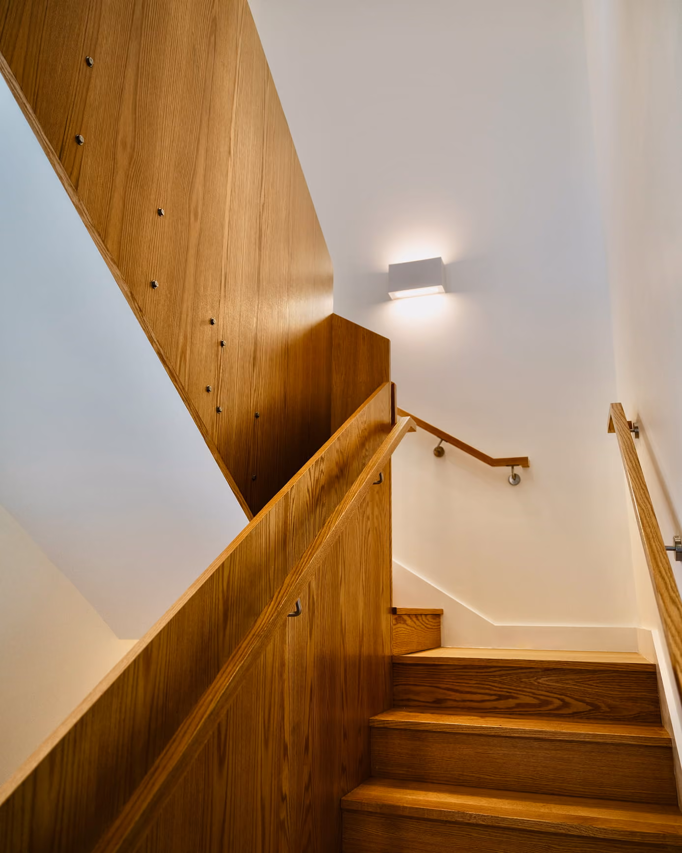 Upward view of a wooden staircase and handrail beneath a wall-mounted light.