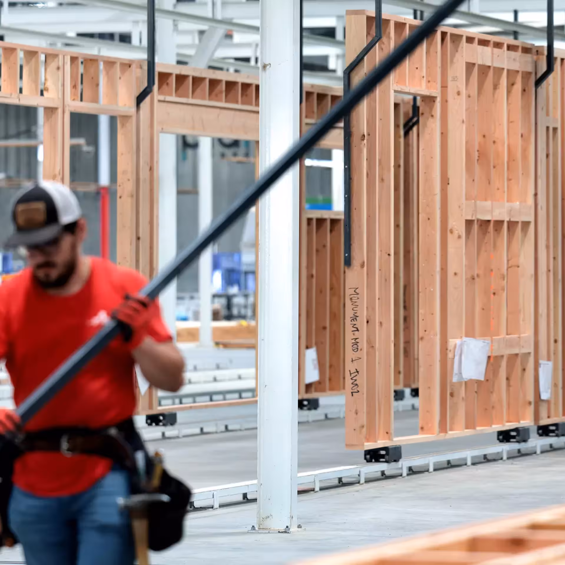 Worker carrying a long black metal beam through a factory where prefabricated wooden wall frames are being assembled on a production line.