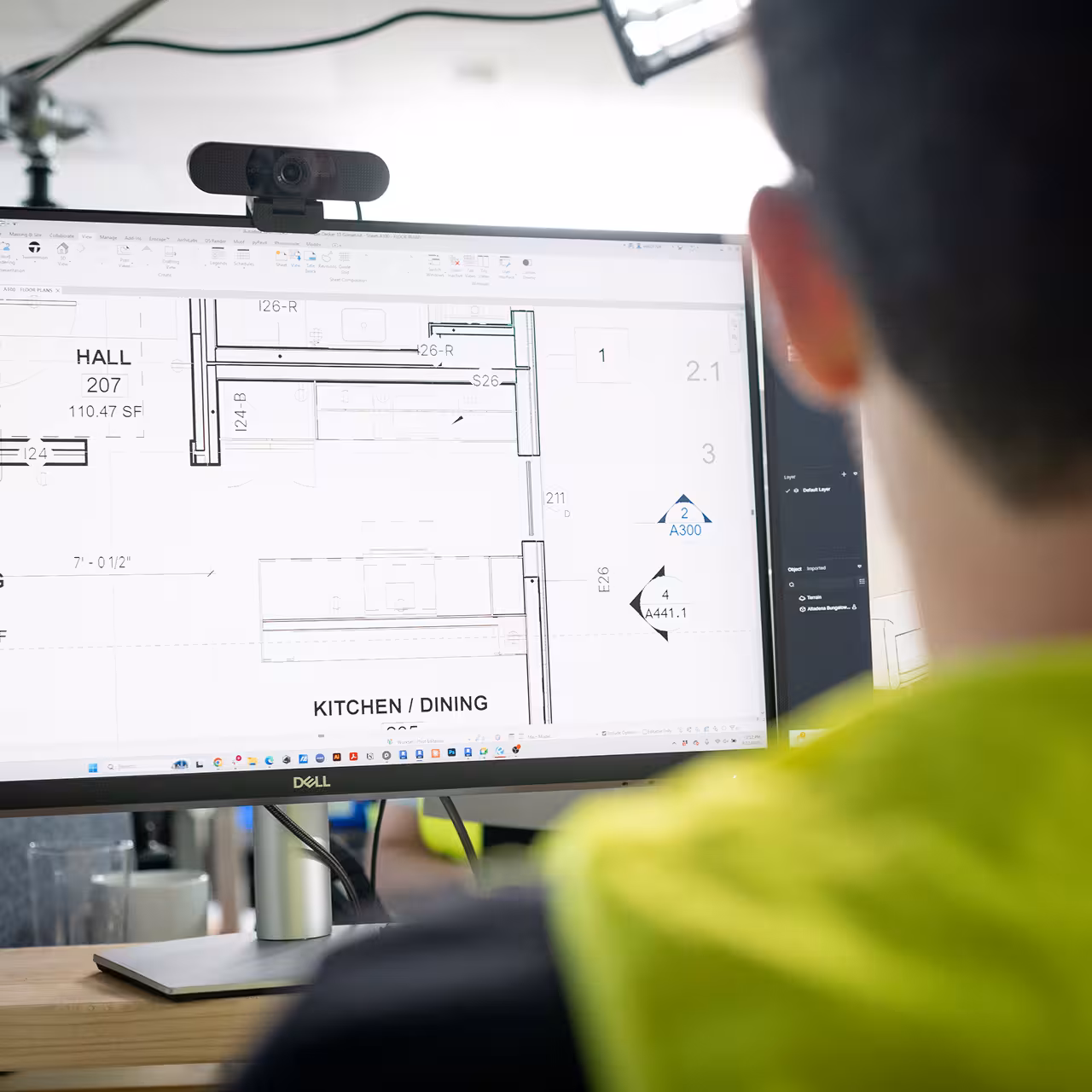 Person in a high-visibility shirt reviewing an architectural floor plan on a large computer monitor in an office.