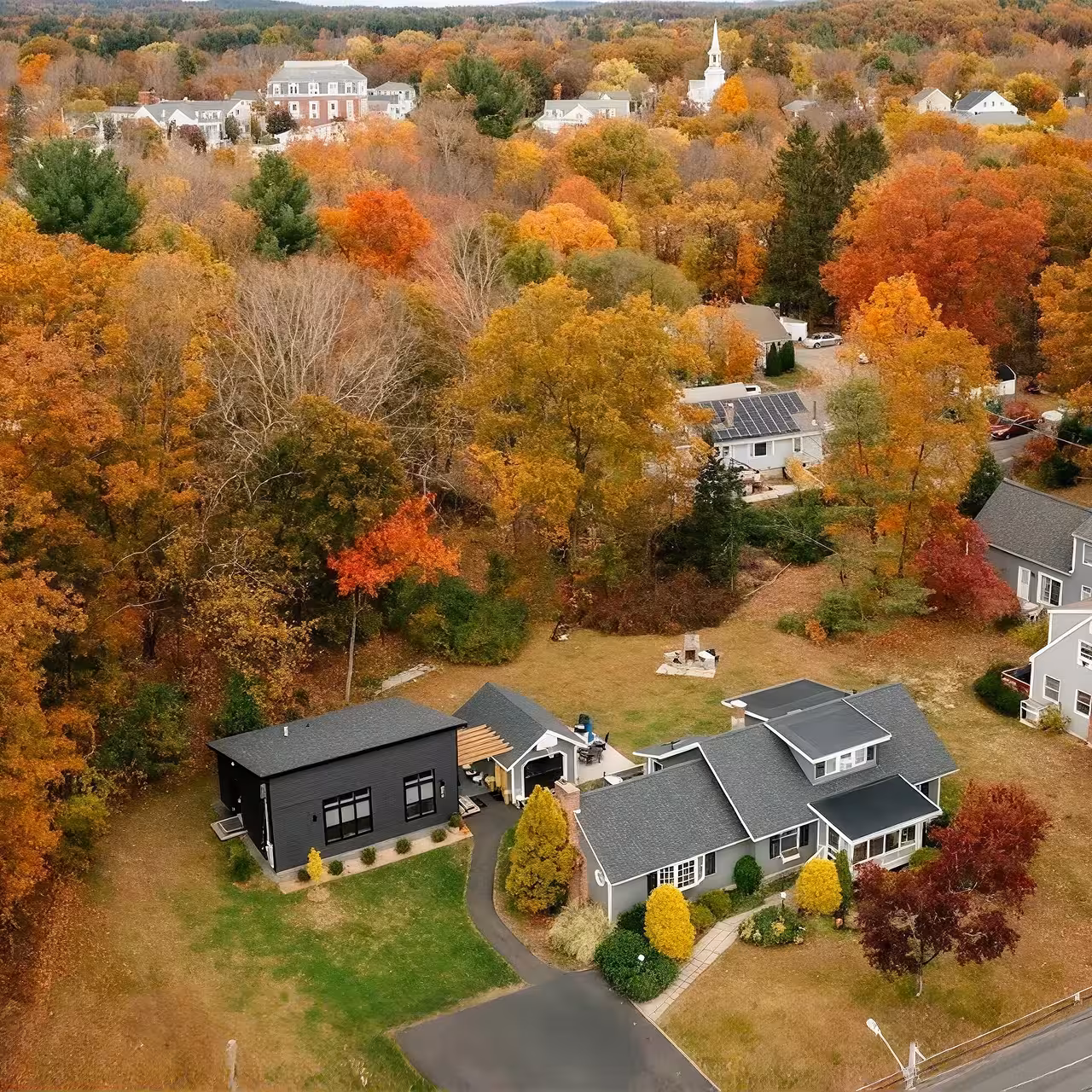 Aerial view of a residential neighborhood surrounded by dense autumn foliage, with detached homes, yards, and a church steeple visible in the distance.