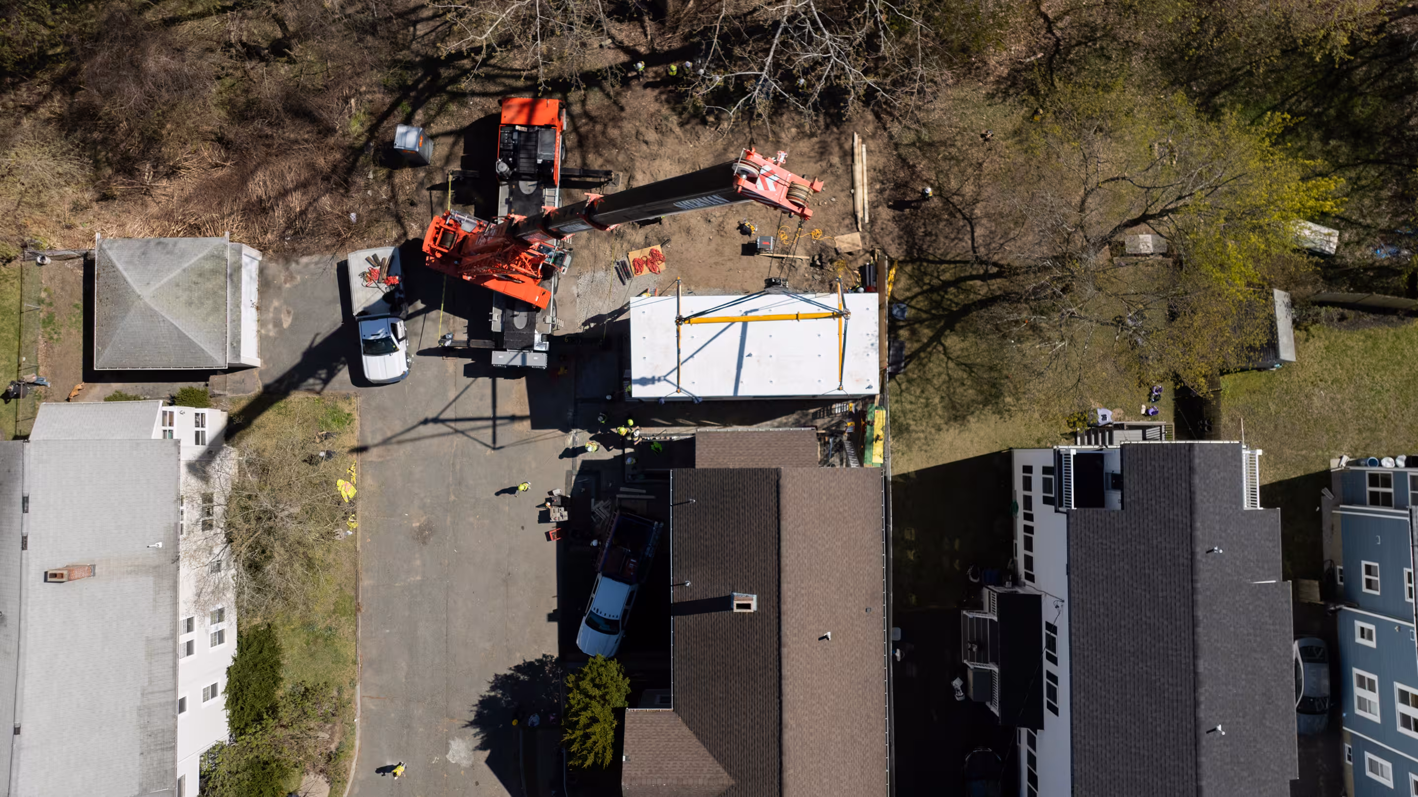 Aerial view of a residential construction site where a large crane is lifting a prefabricated building module into place between neighboring homes.