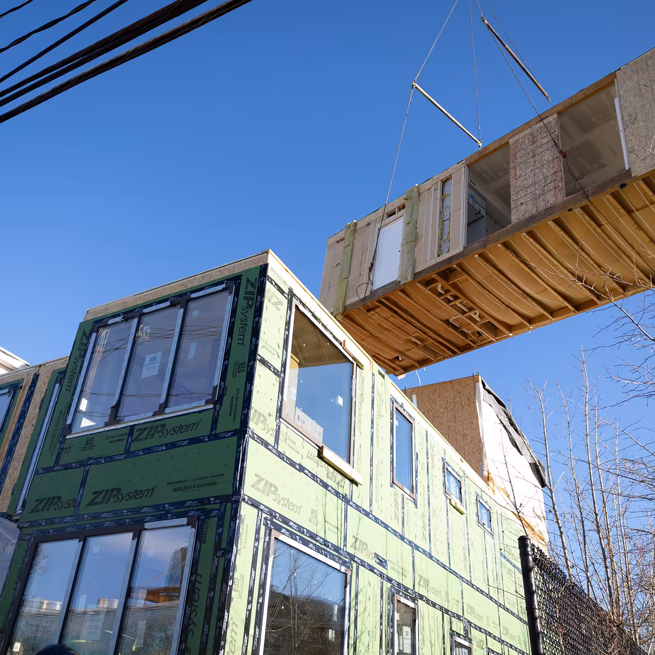 A crane lowers a prefabricated upper-story module onto a partially assembled two-story building wrapped in green exterior sheathing.