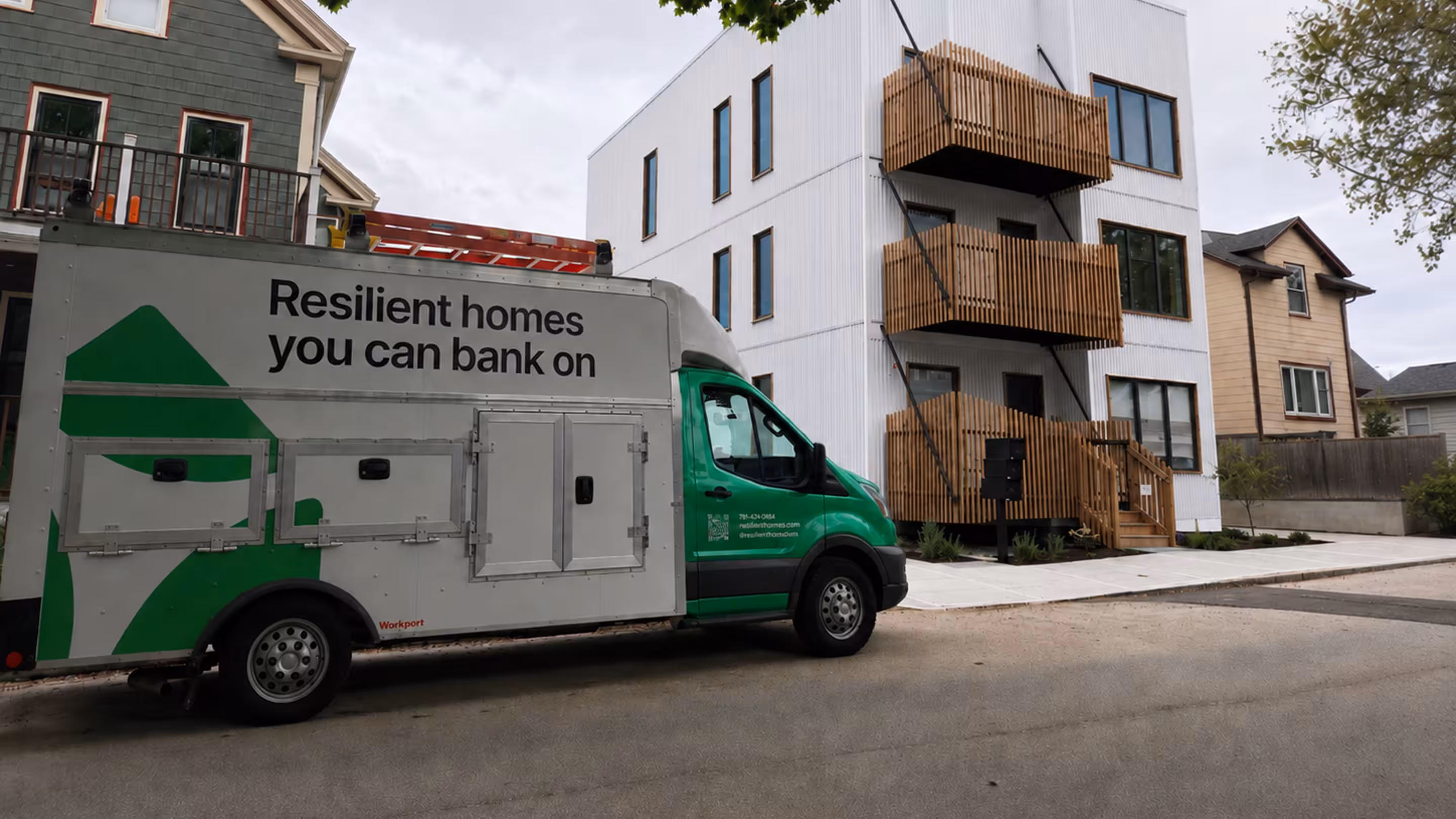 Green and white service truck parked outside a modern three-story residential building with white siding and wooden balconies.
