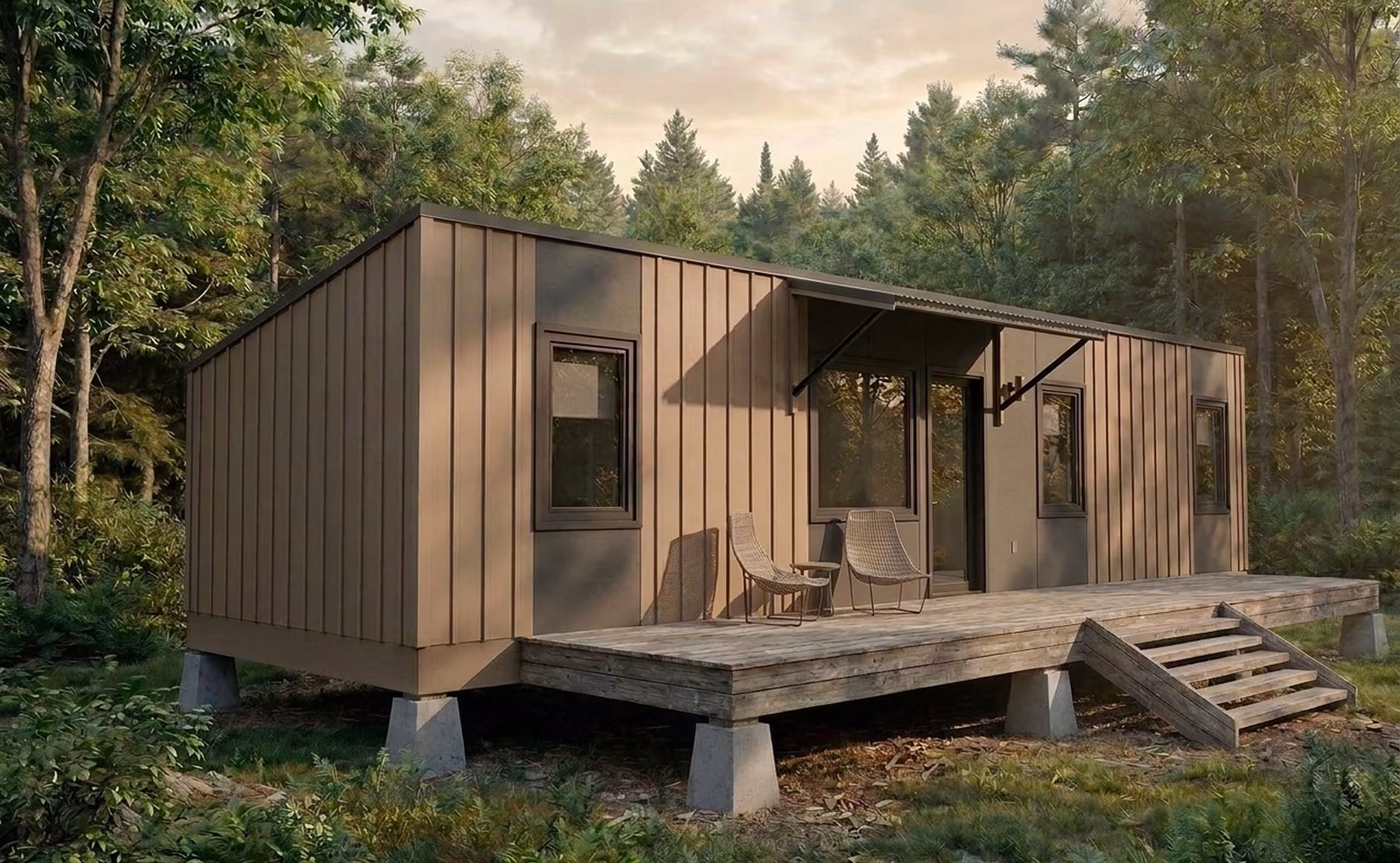 Simple rectangular cabin elevated on concrete piers in a wooded setting, with a wooden deck, outdoor chairs, and entry awning.