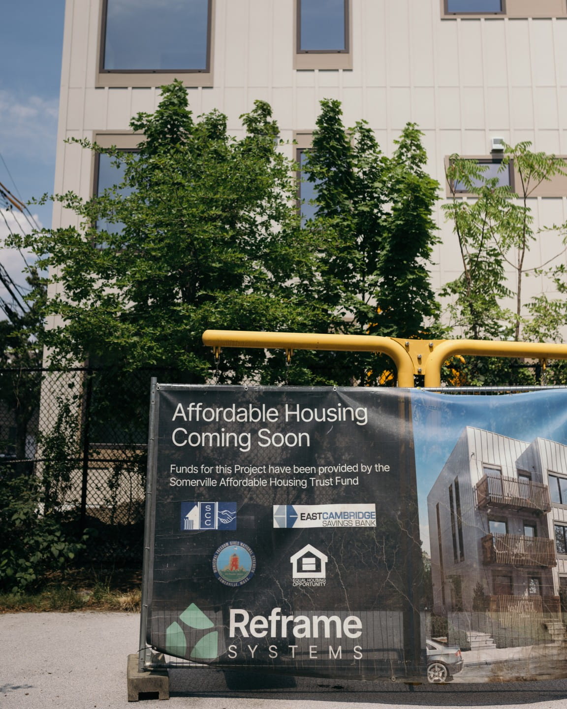 Project information board displayed outside a completed white residential building surrounded by trees.