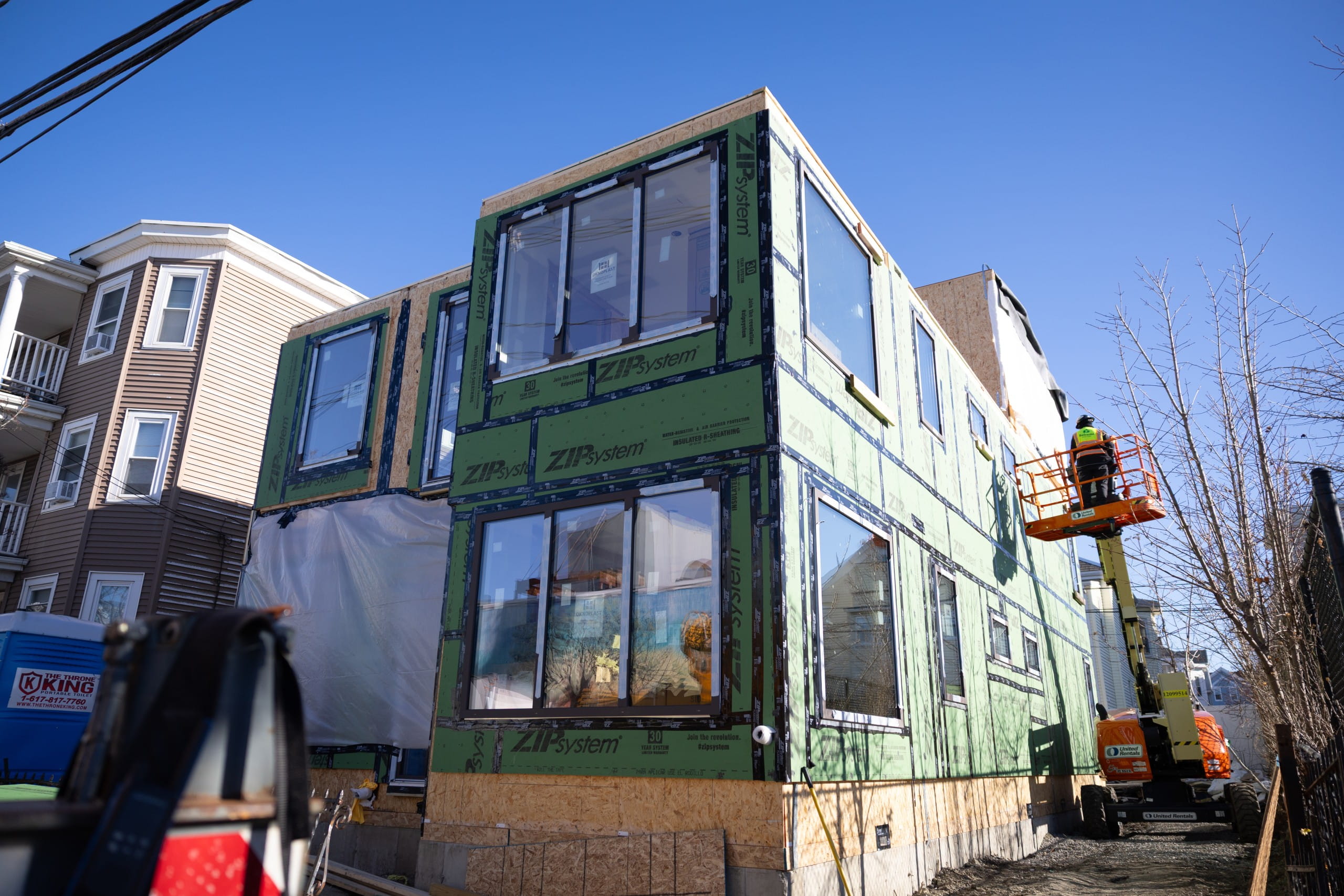 Partially assembled prefabricated building with green exterior sheathing, large windows, and construction equipment nearby.