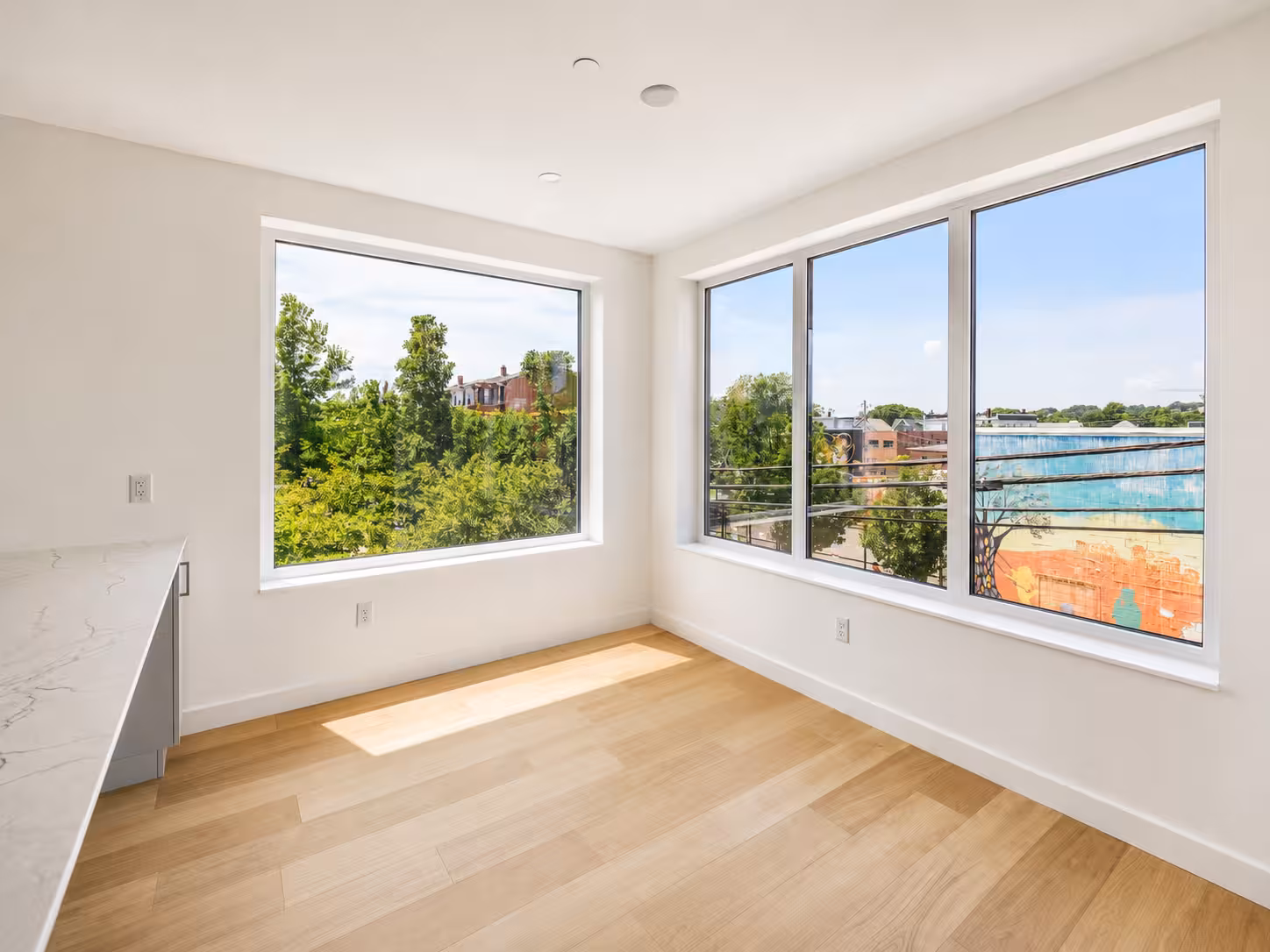 Empty room with light wood floors and large corner windows overlooking trees and surrounding buildings.