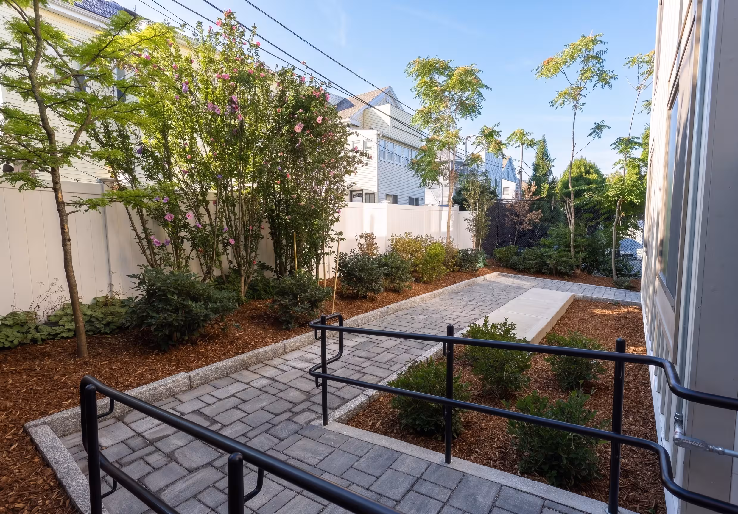 Outdoor walkway bordered by trees, planted garden beds, and black metal railings beside the building.