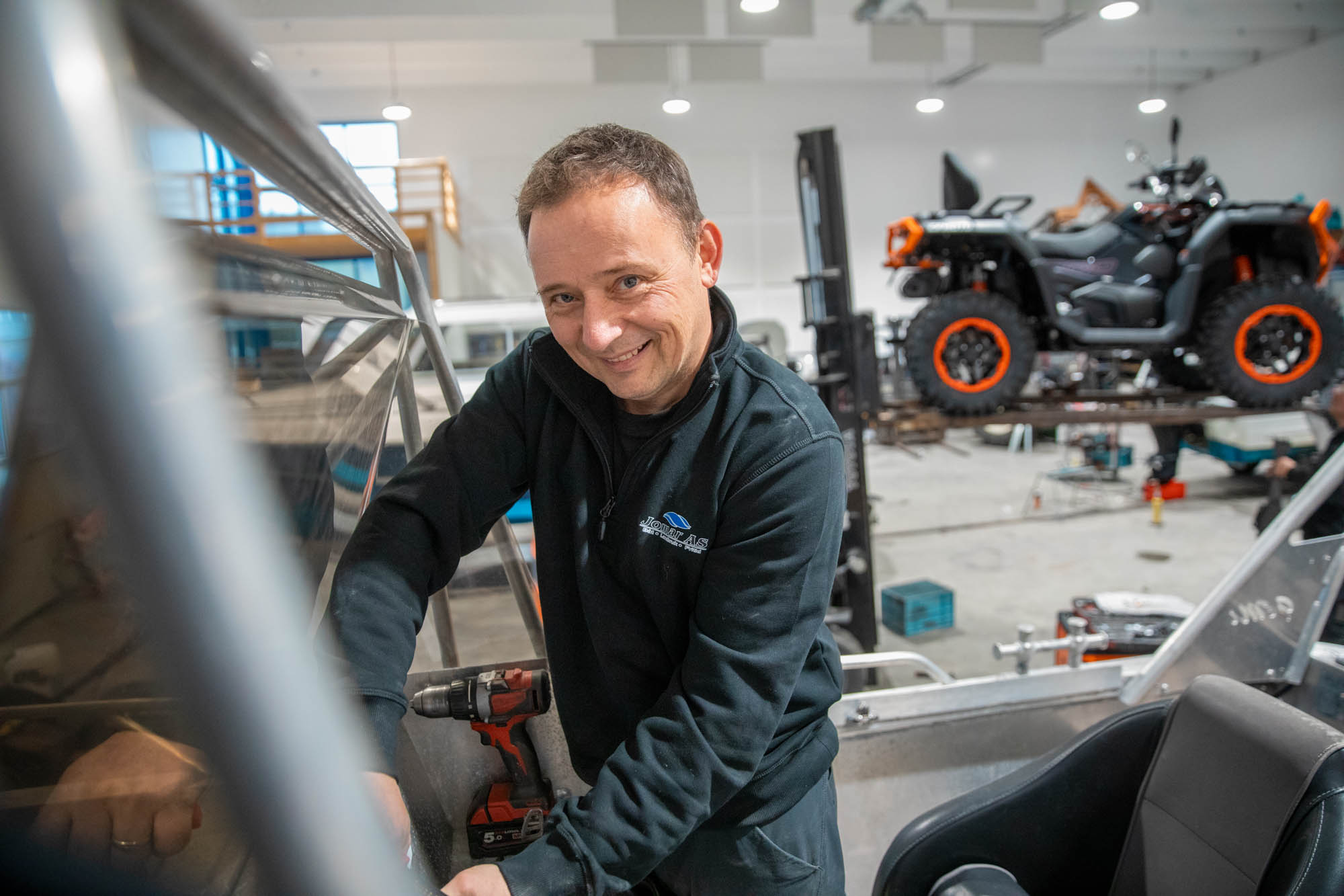 Mann smiler mens han arbeider med en rød og svart drill i et verksted med et terrengkjøretøy i bakgrunnen.