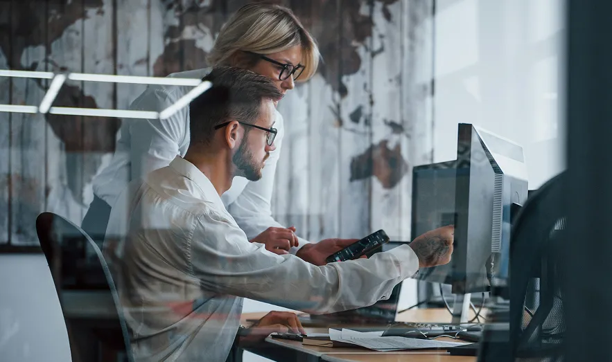 Image of two professionals collaborating at a desk in a modern office space, with one person pointing at a computer screen while the other observes, reflecting teamwork and innovation.