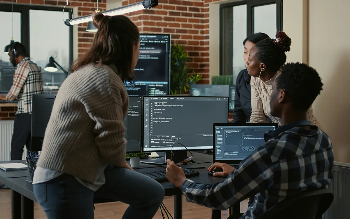 Image of a diverse team of professionals collaborating in a modern office, working together on coding tasks with multiple screens displaying code and software development tools.