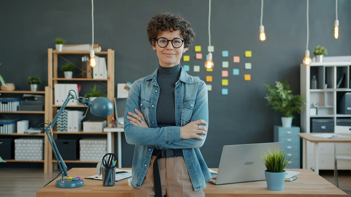 Portrait of a professional woman with curly hair, wearing glasses and a denim jacket, standing confidently in an office space with a modern, creative workspace backdrop.