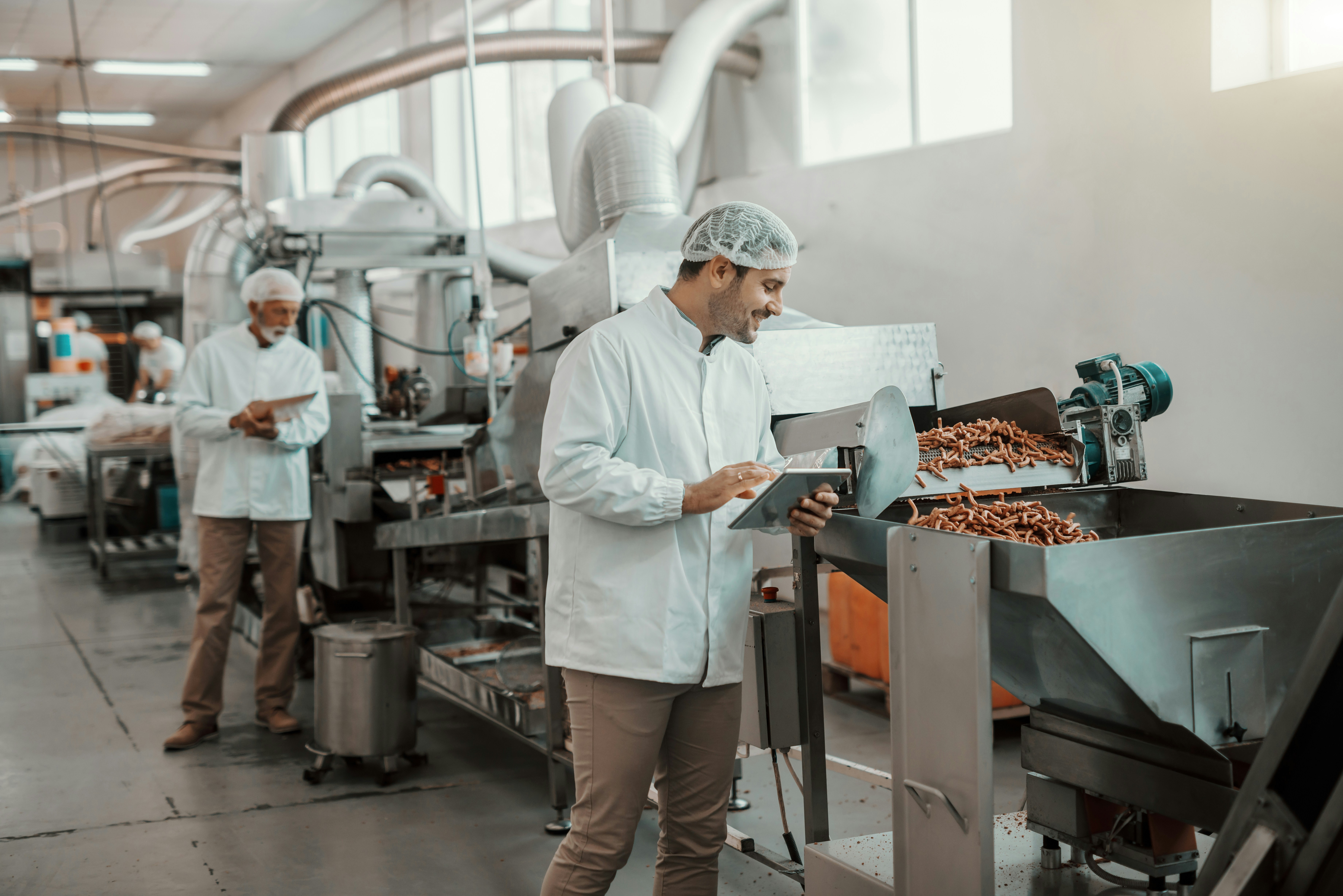 Deux hommes en blouse surveillent une machine traitant des aliments dans une usine agroalimentaire.