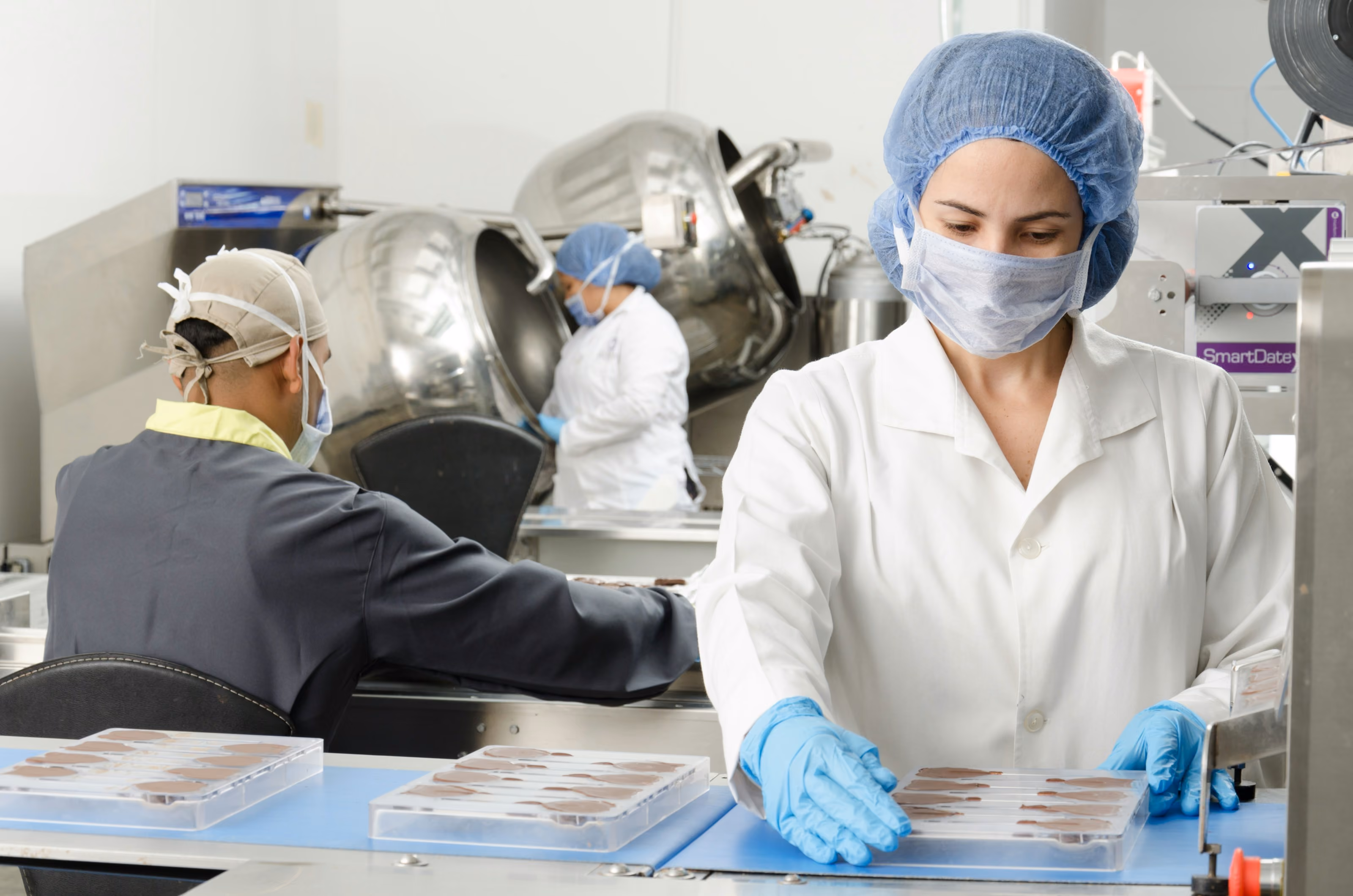 Travailleurs en blouse et masque manipulant des moules à chocolat dans une usine alimentaire.