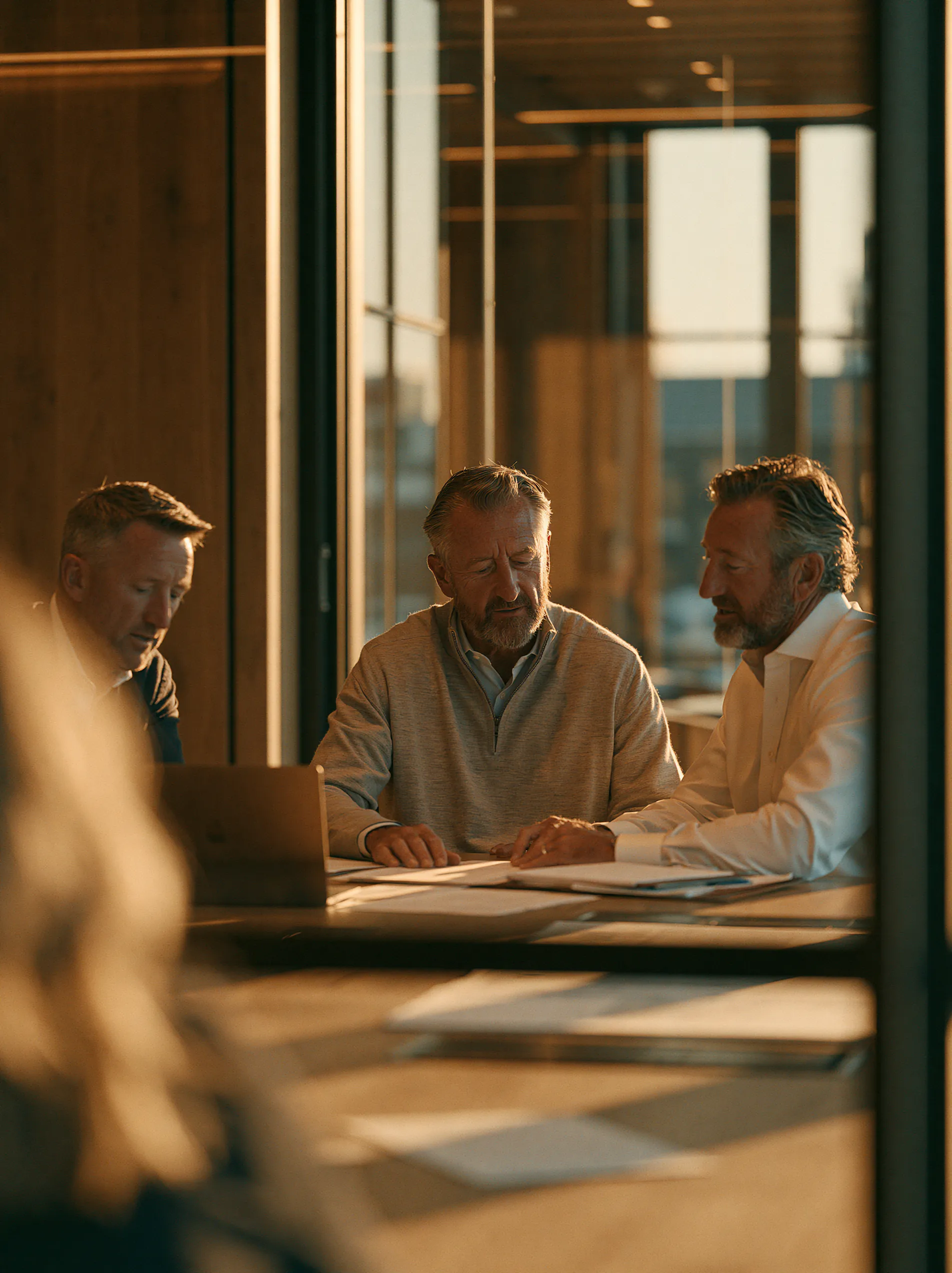 Réunion de travail entre trois dirigeants dans une salle de réunion moderne baignée par une lumière chaude de fin de journée. Les participants examinent des documents et échangent autour d’un ordinateur portable dans un environnement premium aux finitions en bois et en verre.