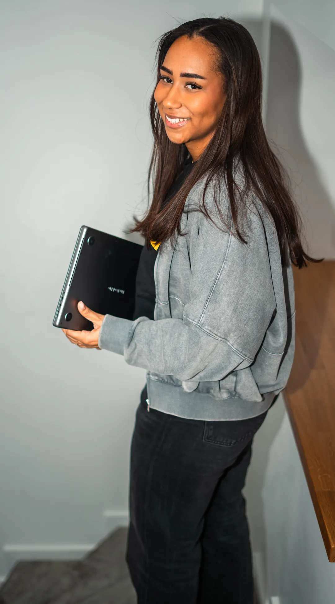 Smiling person holding a laptop and looking toward the camera in an indoor setting.