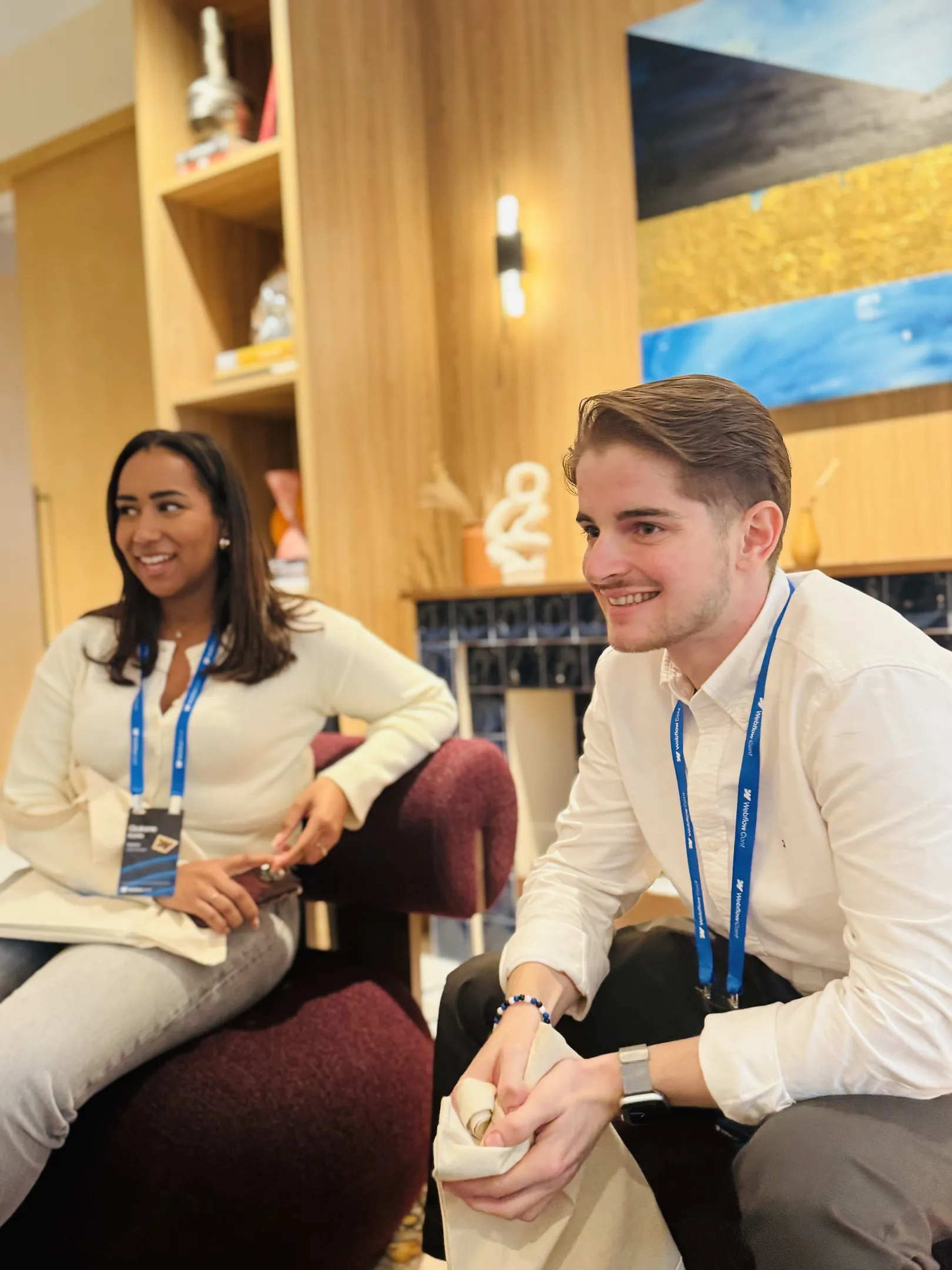 Deux personnes portant des cordons de conférence assises et souriantes dans une pièce confortable et moderne avec des étagères en bois et des œuvres d'art colorées.