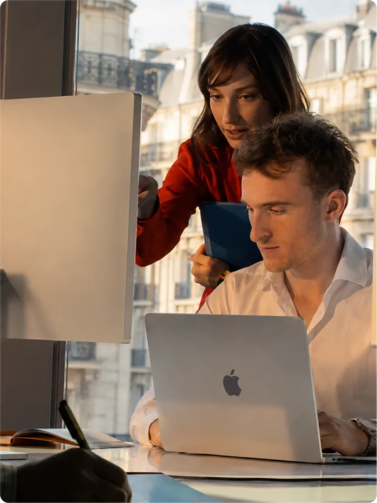AI-generated image showing two professionals collaborating in an office, reviewing a website on a laptop with Parisian Haussmann buildings visible through the window.