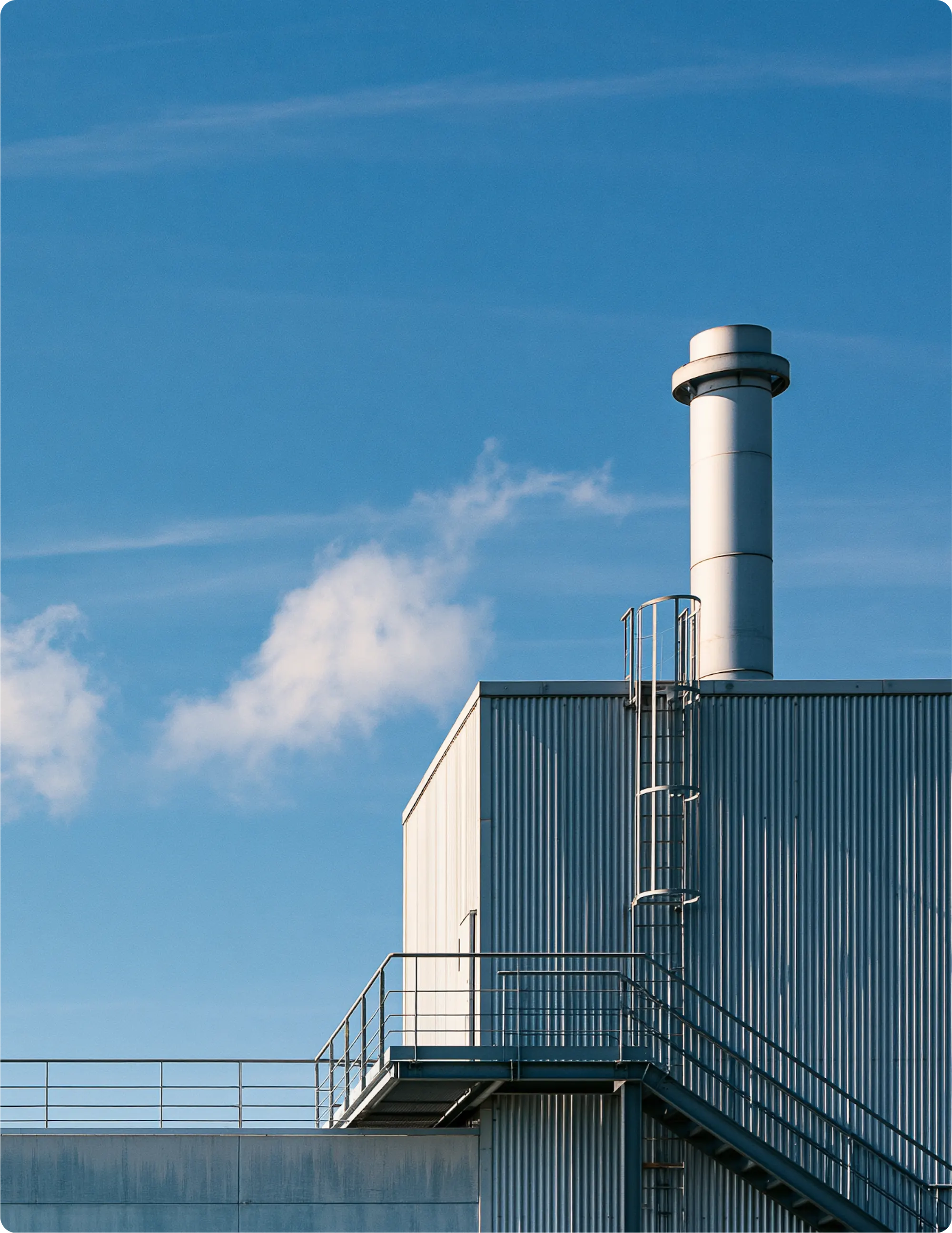 Bâtiment industriel avec cheminée métallique, escaliers extérieurs et balustrade sur fond de ciel bleu clair.