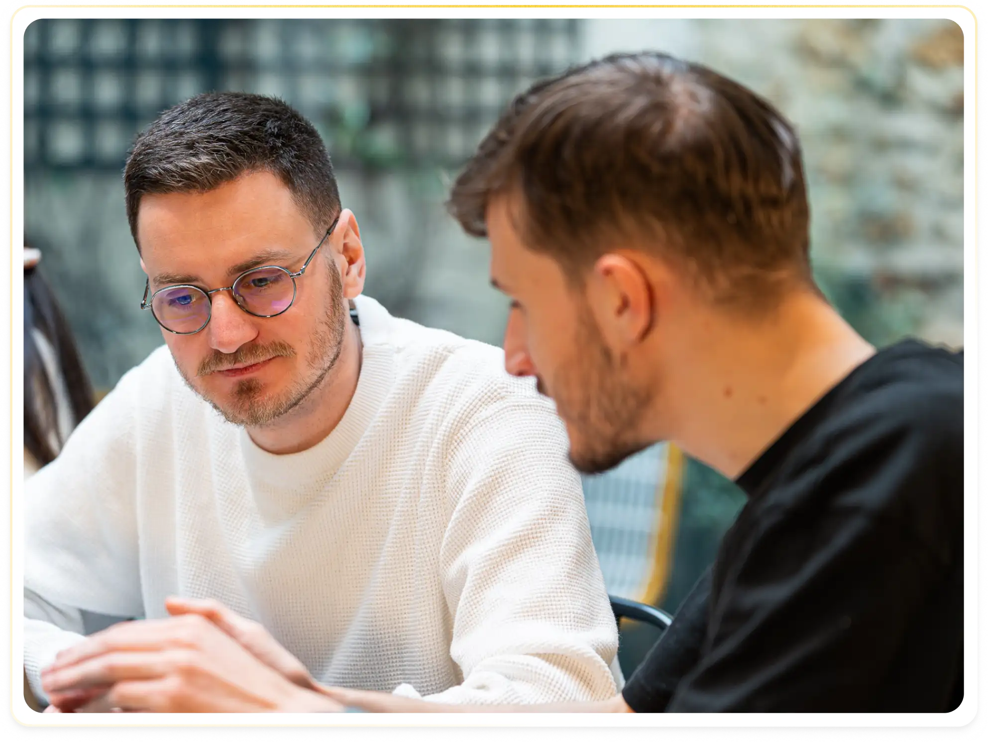 Two men focused on a discussion, one wearing glasses and a white sweater, the other in a black shirt.