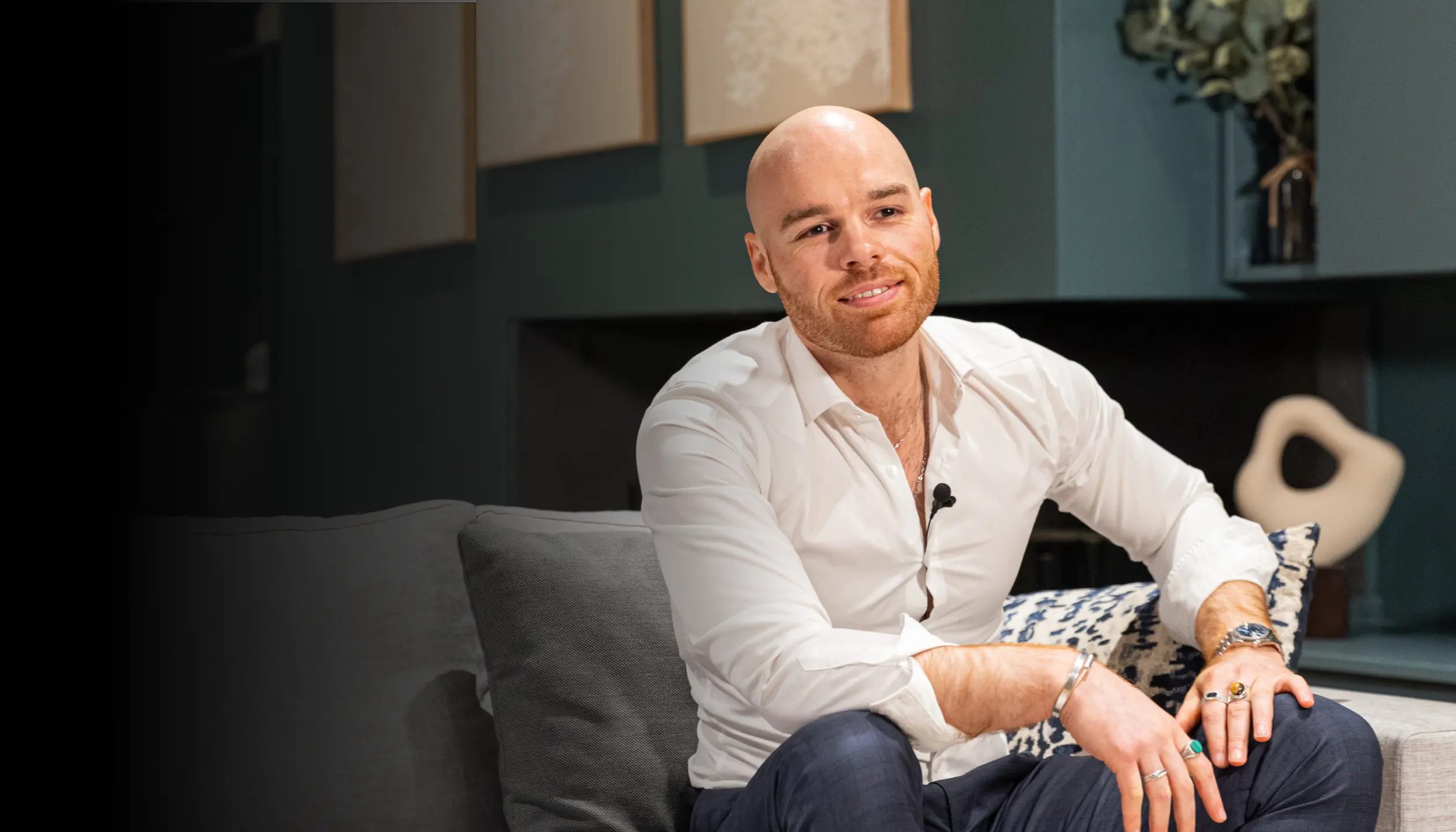 A smiling man sitting on a couch, wearing a white shirt and a lavalier microphone. The setting is stylish and cozy, with the Avant Garde Groupe and Digidop logos displayed in the bottom left corner of the image.