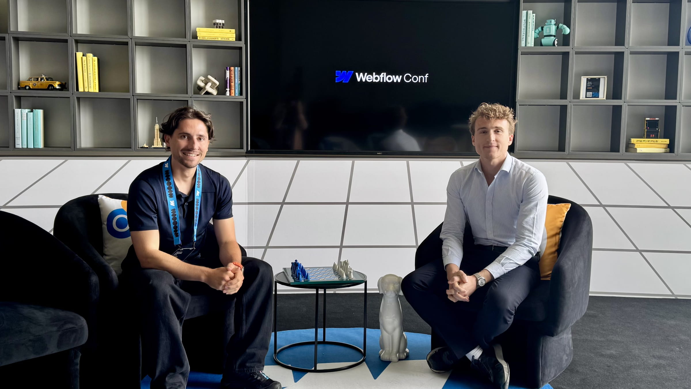 Thomas Labonne (à gauche) et Florian Bodelot (à droite) assis dans un salon lors de la Webflow Conf, devant un écran affichant le logo Webflow Conf, dans un décor moderne avec étagères et objets design.