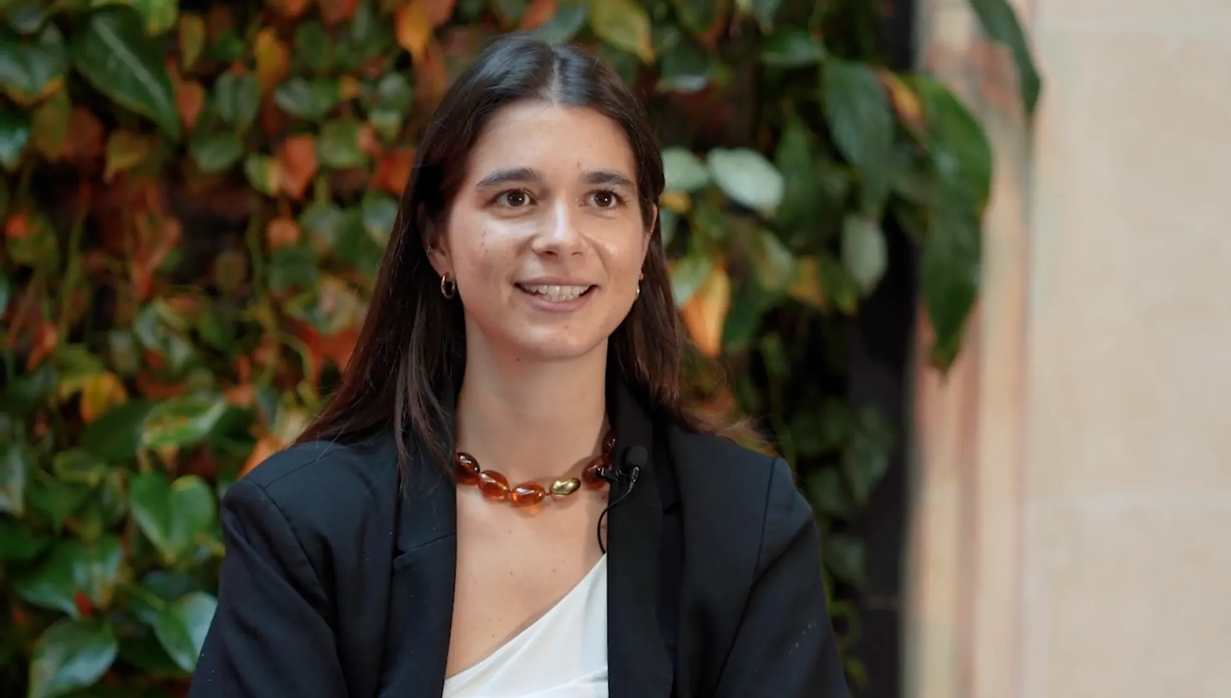 Portrait d’une femme en entretien, portant un blazer noir et un collier en résine, avec un mur végétal en arrière-plan.
