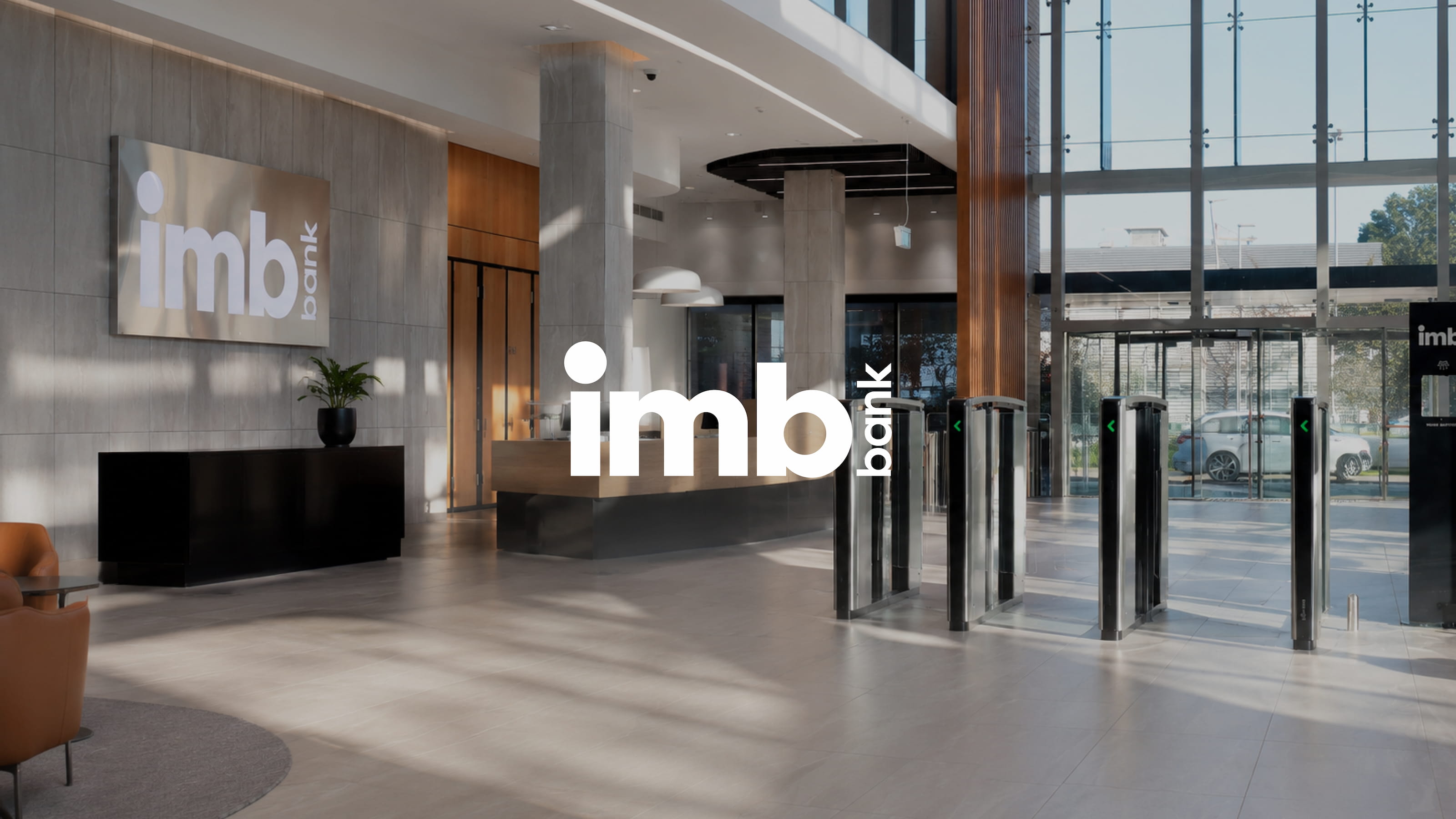 Modern reception area of an IMB Bank branch featuring a spacious double-height lobby, floor-to-ceiling glass façade, contemporary security gates, natural daylight, and prominent IMB Bank branding.