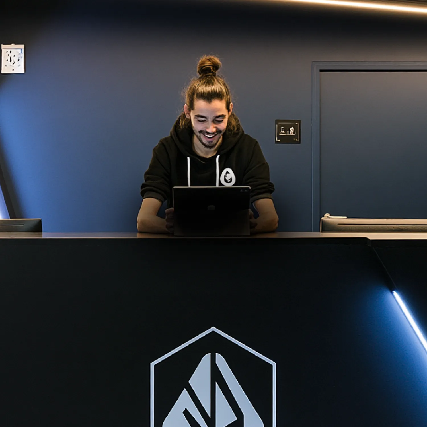 Employé d’un centre de fitness utilisant une tablette derrière le comptoir d’accueil d’une salle de sport moderne, dans un environnement minimaliste aux éclairages LED.