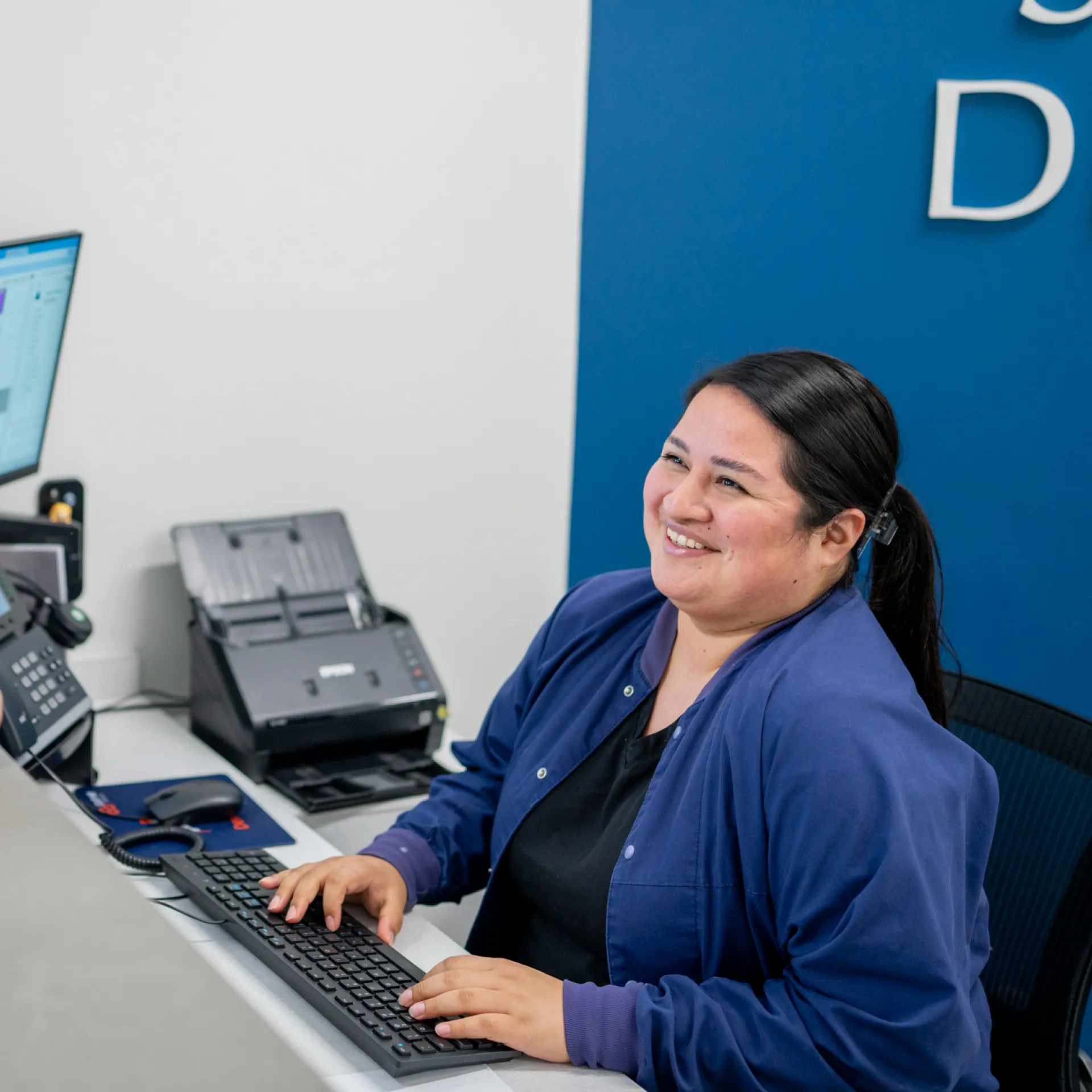 Office photo: Person with dark hair, smiling, in a blue jacket, working at a desk with a computer and phone.