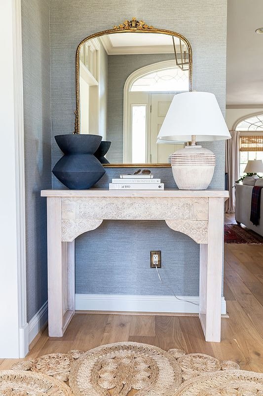 A pale carved console table against a wall with a lamp and vases on top.
