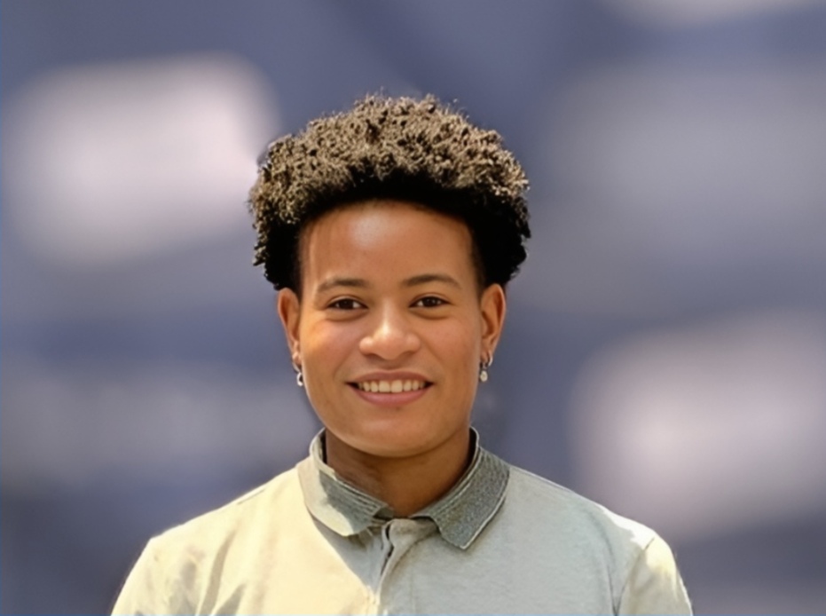 A person with light brown skin and a medium length afro adorns silver small hoop earrings and a grey collared shirt. 