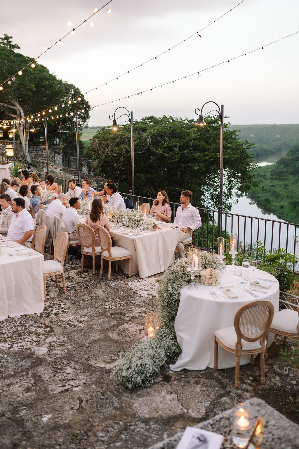 Outdoor evening wedding reception with guests seated at decorated tables covered with white linens and floral arrangements, overlooking a river and lush greenery, illuminated by string lights and candles.