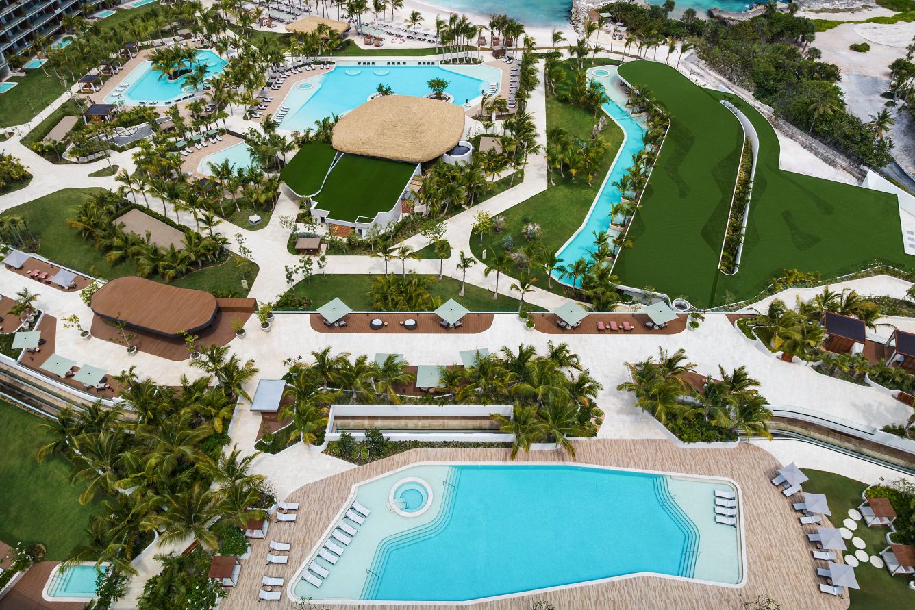 Aerial view of a luxury resort featuring multiple swimming pools surrounded by lounge chairs, palm trees, green lawns, and pathways, with a thatched-roof building and a mini golf course near the coast.