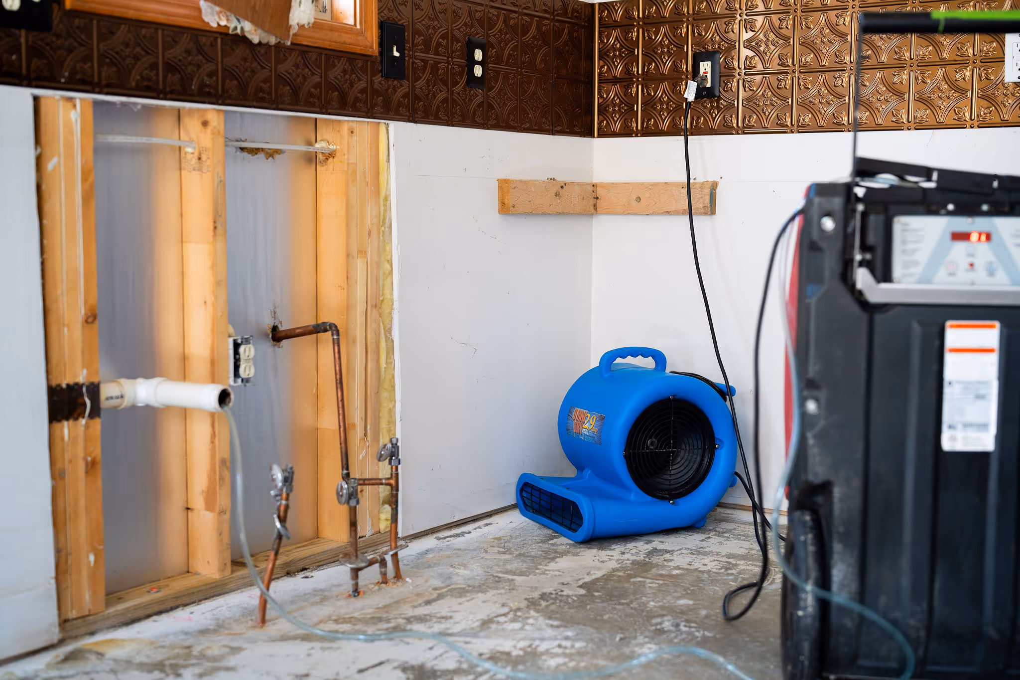 A blue air mover dries out a wall that's been cut open to reveal pipes underneath copper tile.
