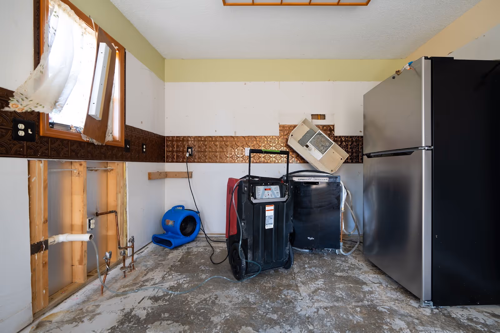 A partially restored kitchen with exposed pipes, a damaged fridge, and drying equipment.