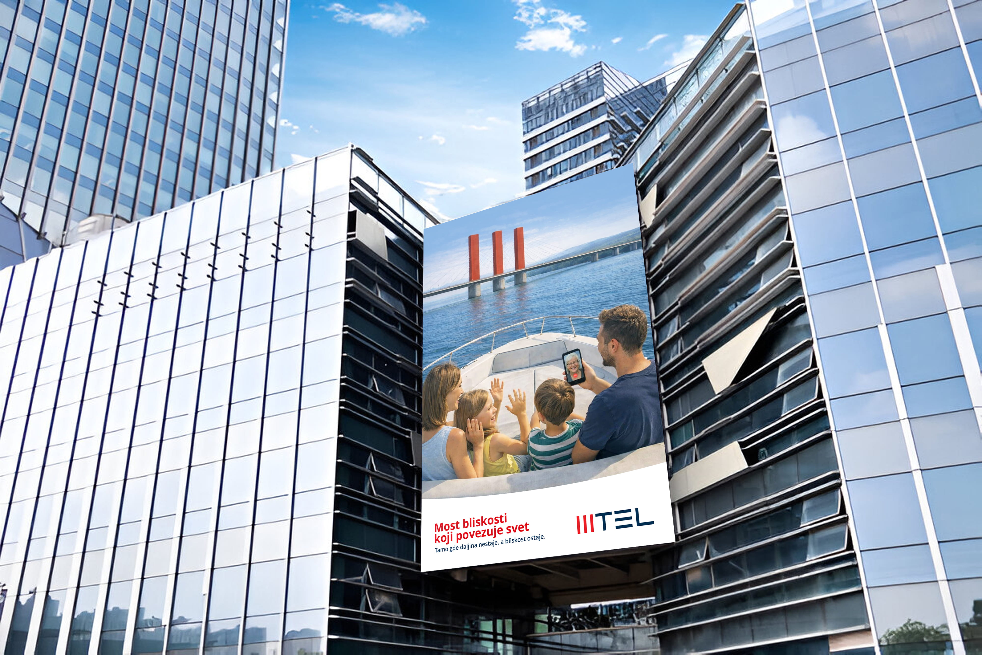 Billboard on glass building shows family on boat waving to person on a phone video call with a bridge view.