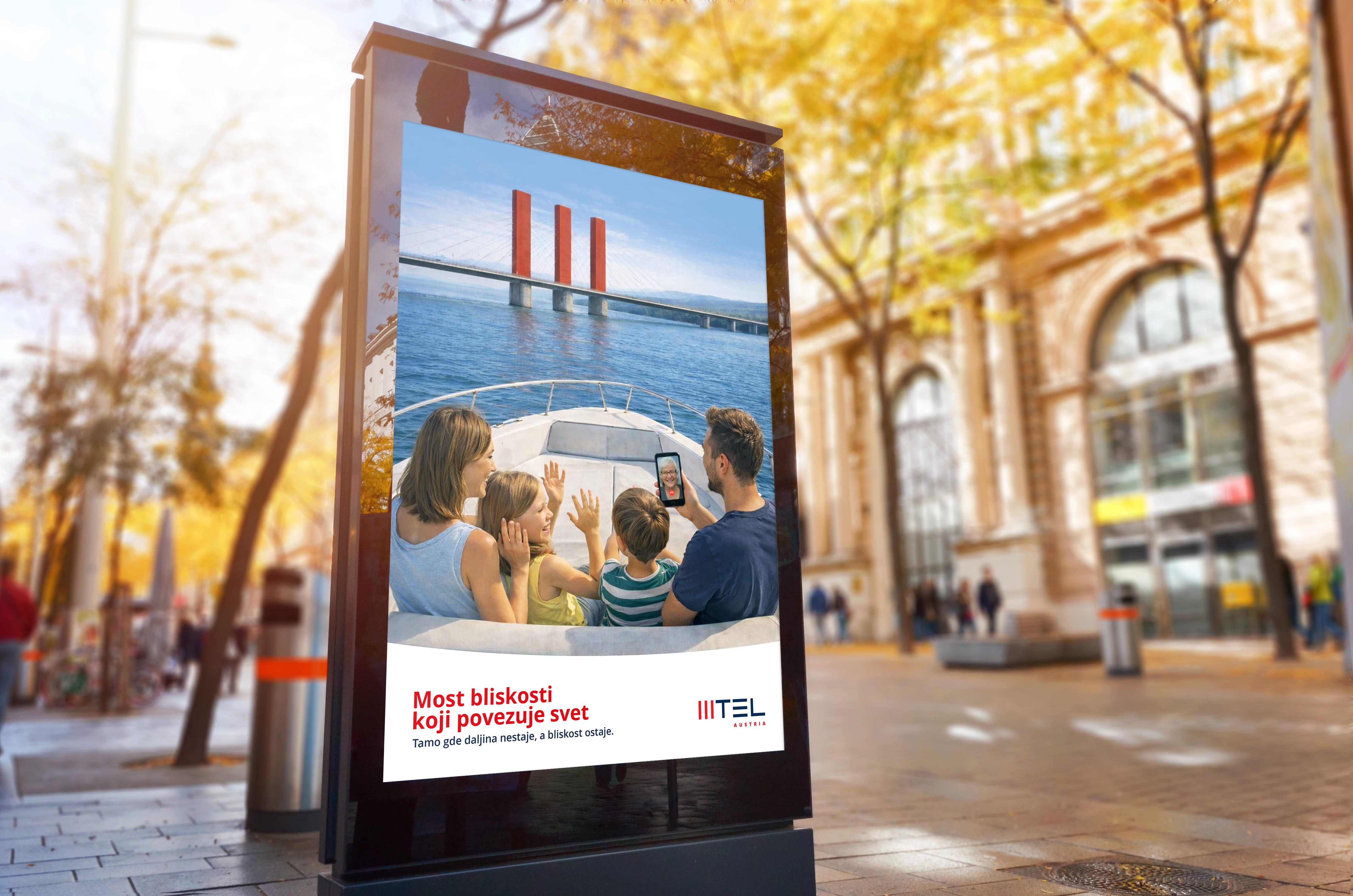 Family on boat waving during video call with a person on phone, near a bridge over water.