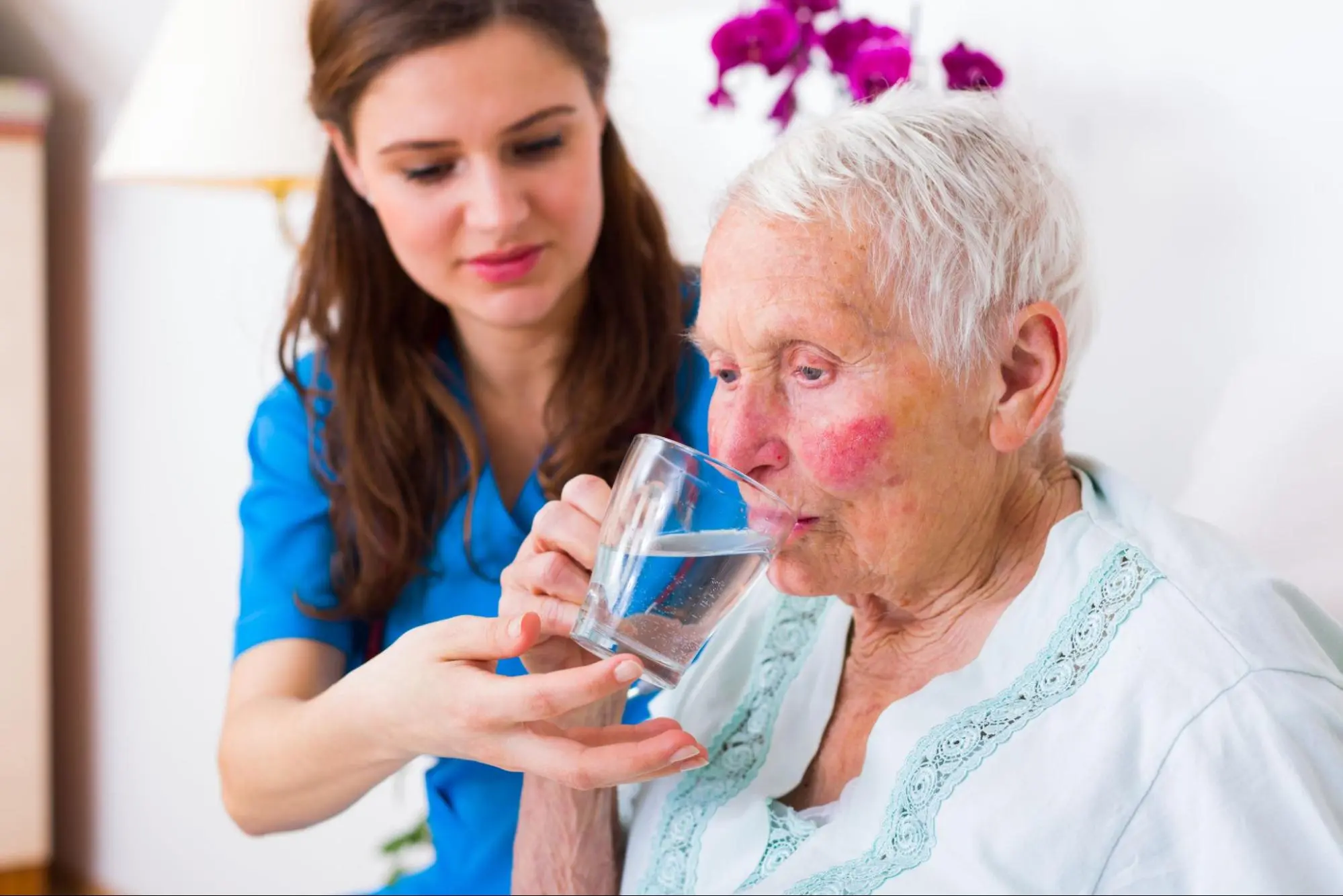 Female caregiver in blue scrubs helping an elderly senior woman drink a glass of water.