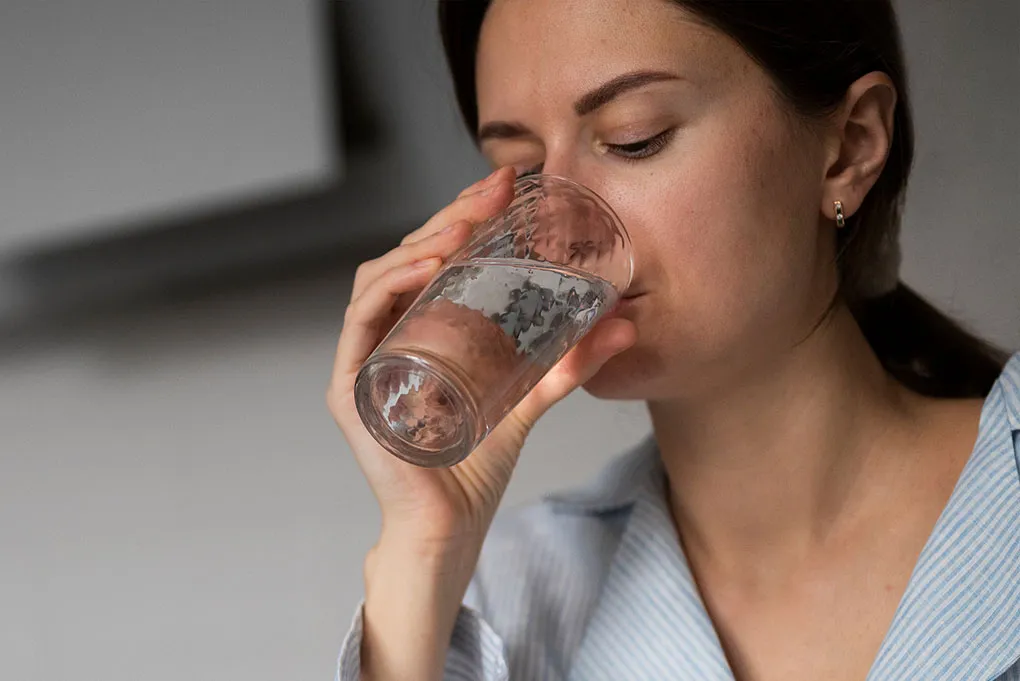 A woman with her eyes closed taking a sip from a clear glass of water.