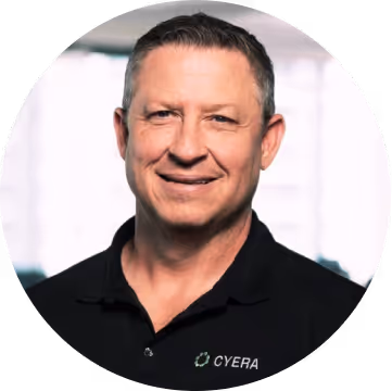 Man with short dark hair smiling, wearing a black shirt with a Cyera logo, in front of a blurred bright window.