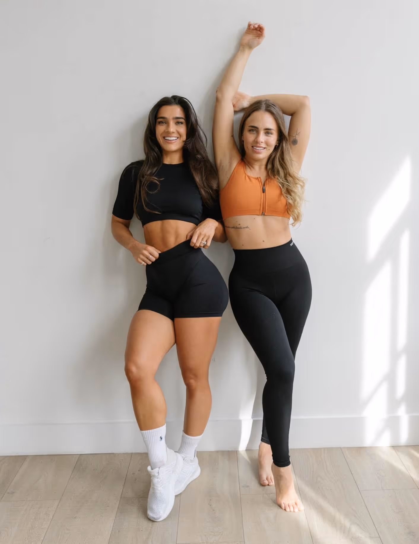 Two fit women smiling and posing against a white wall wearing black workout bottoms and crop tops, one barefoot and the other in white sneakers.