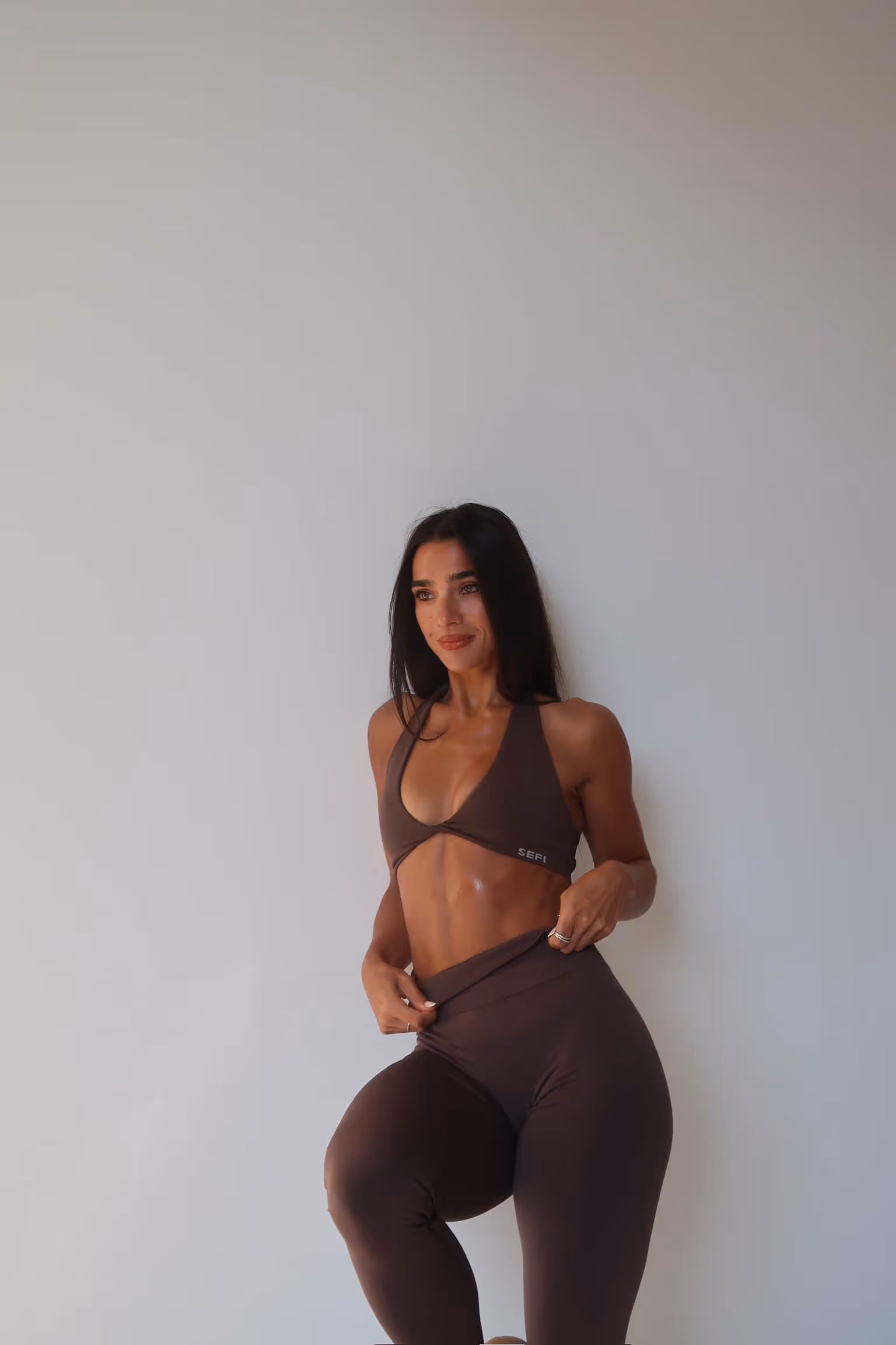 Fit woman with long dark hair wearing a brown sports bra and leggings standing against a plain light-colored wall.