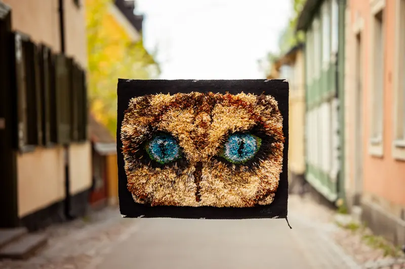 Väggtextil med ugglemotiv och turkosa ögon, fotograferad utomhus på en gata.