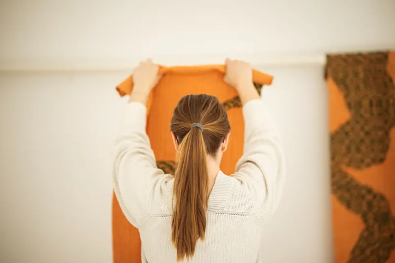 En person hänger upp en orange vävd textil på en ljus vägg.