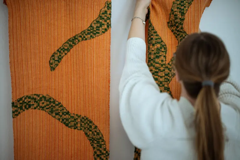 En person justerar en orange väggtextil med gröna slingrande former.